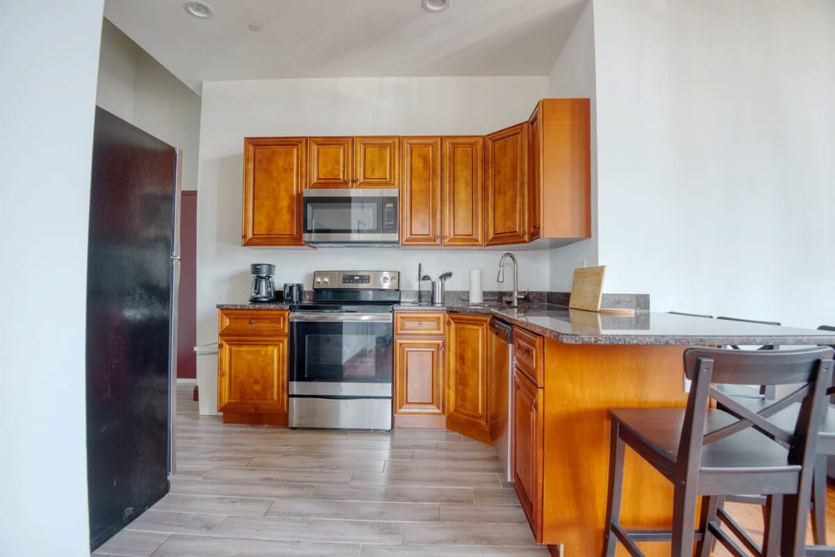Functional kitchen with warm wooden cabinetry, dark granite countertops, and stainless steel appliances including an oven, microwave, and dishwasher. A breakfast bar with dark stools extends into the living space, set against a light tiled floor.