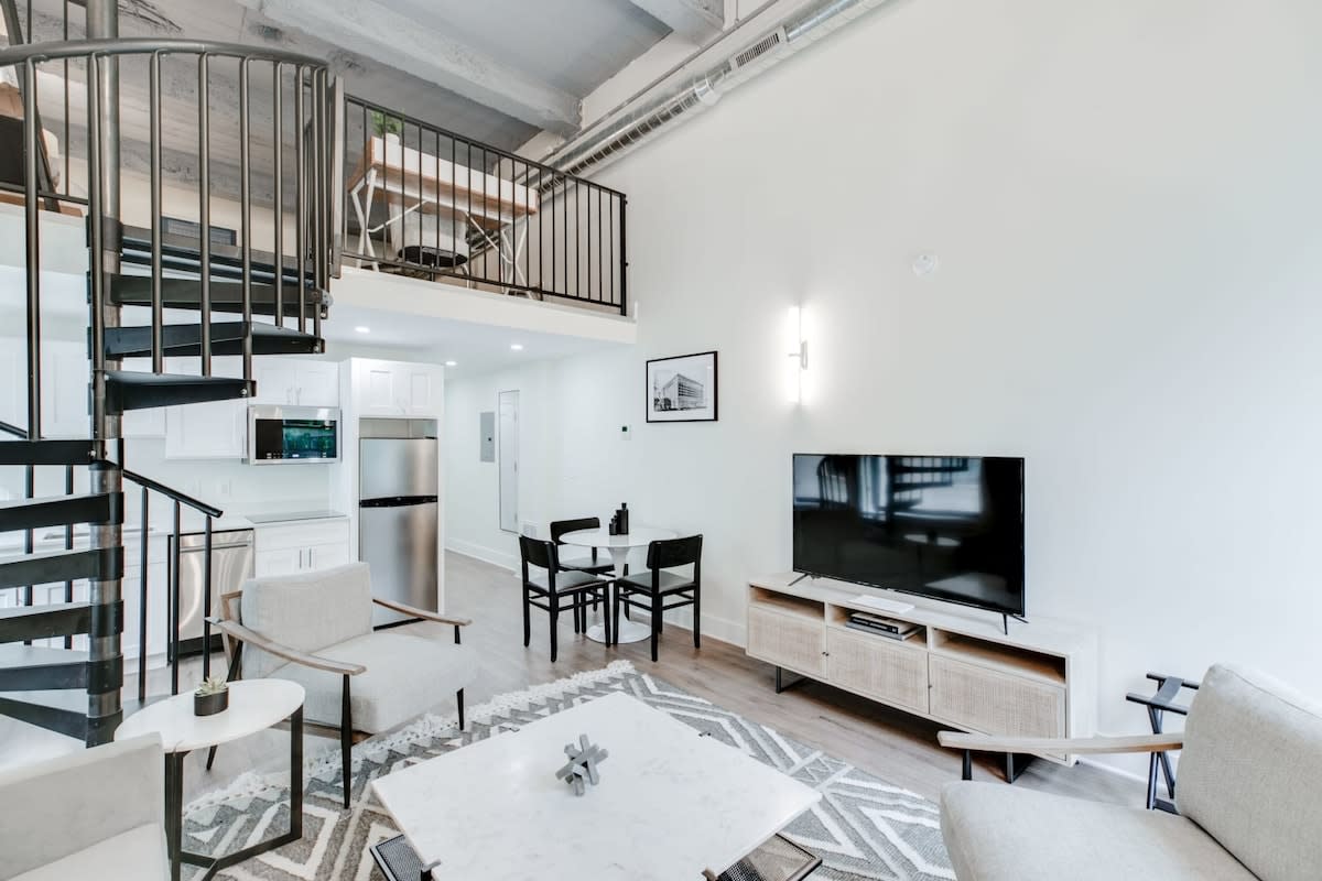 A bright, open-concept loft space featuring a living area with a TV on a console, a light gray sofa, and armchairs, adjacent to a compact white kitchen, all connected by a black spiral staircase leading to a mezzanine level.