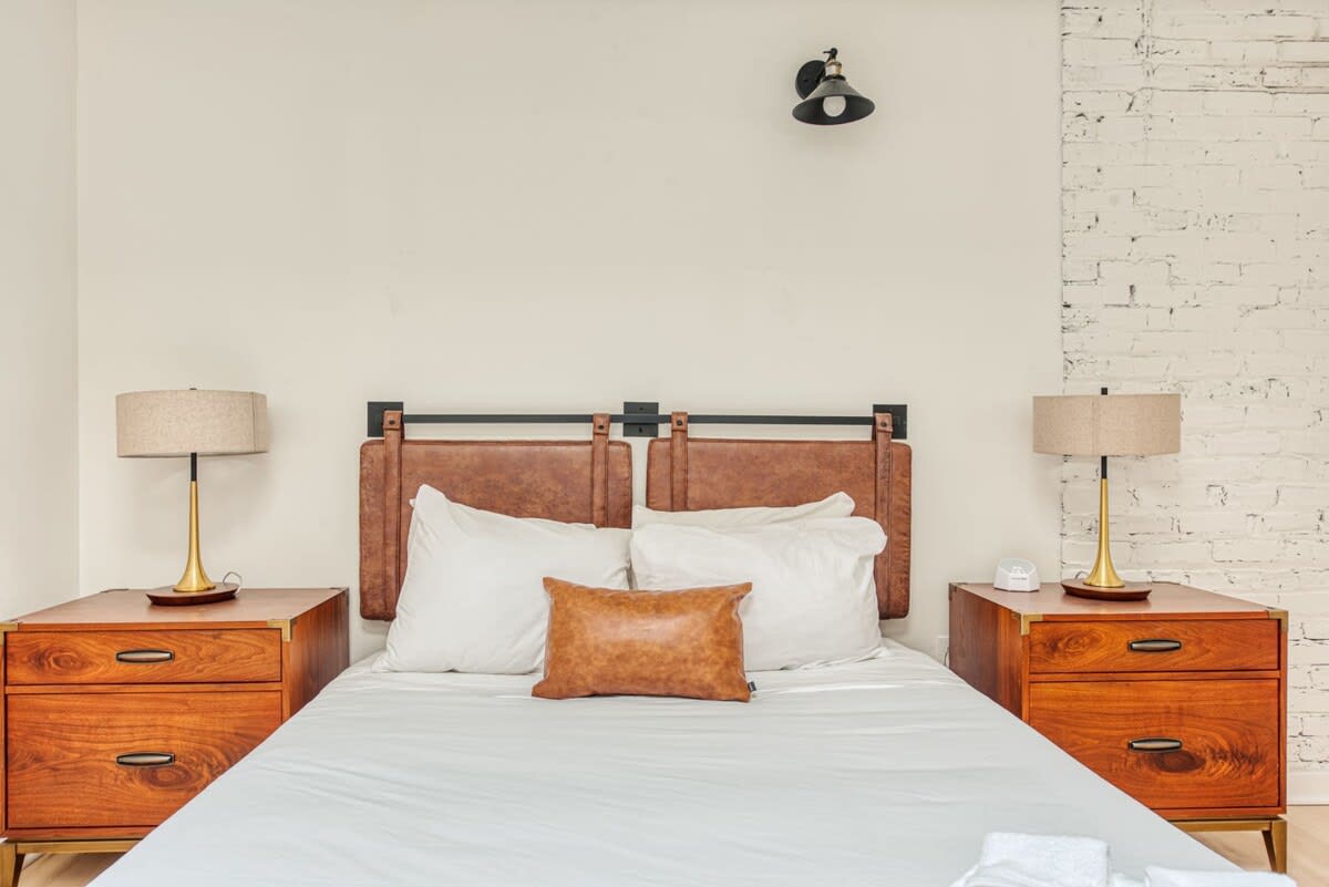 Bedroom with a queen-sized bed featuring a distinctive brown leather headboard, flanked by two wooden nightstands with gold-based lamps, against a light wall with exposed white brick on the right.