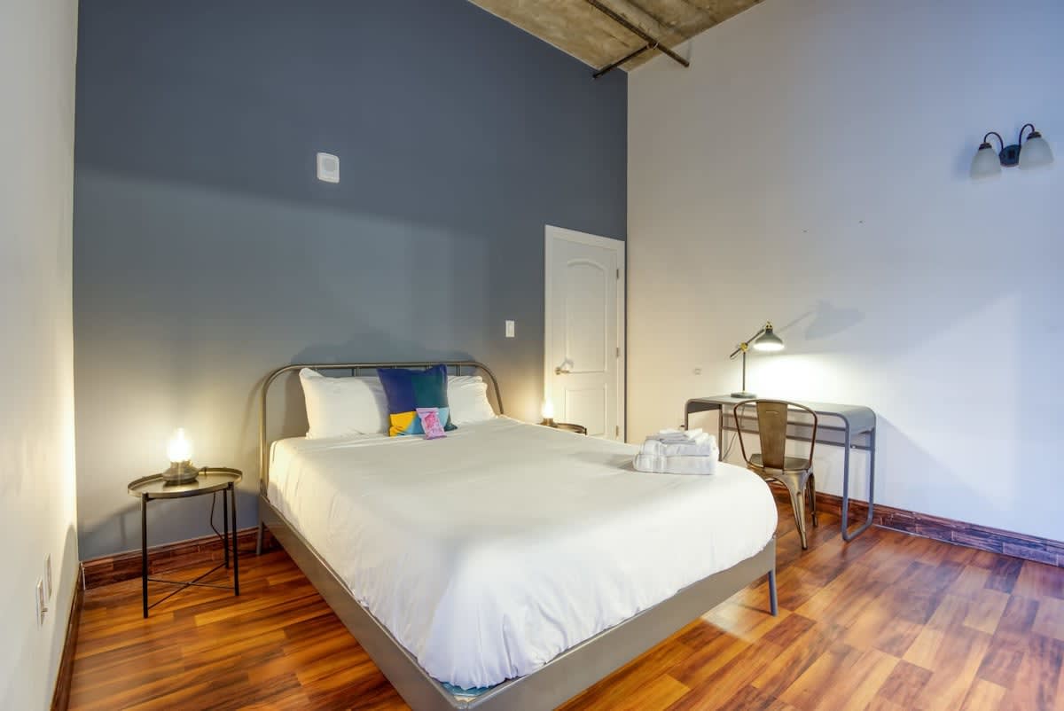 A minimalist bedroom with a queen-sized bed featuring a metal frame and white bedding, flanked by two round metal nightstands with lamps, and a metal desk with a task lamp against a two-tone painted wall.
