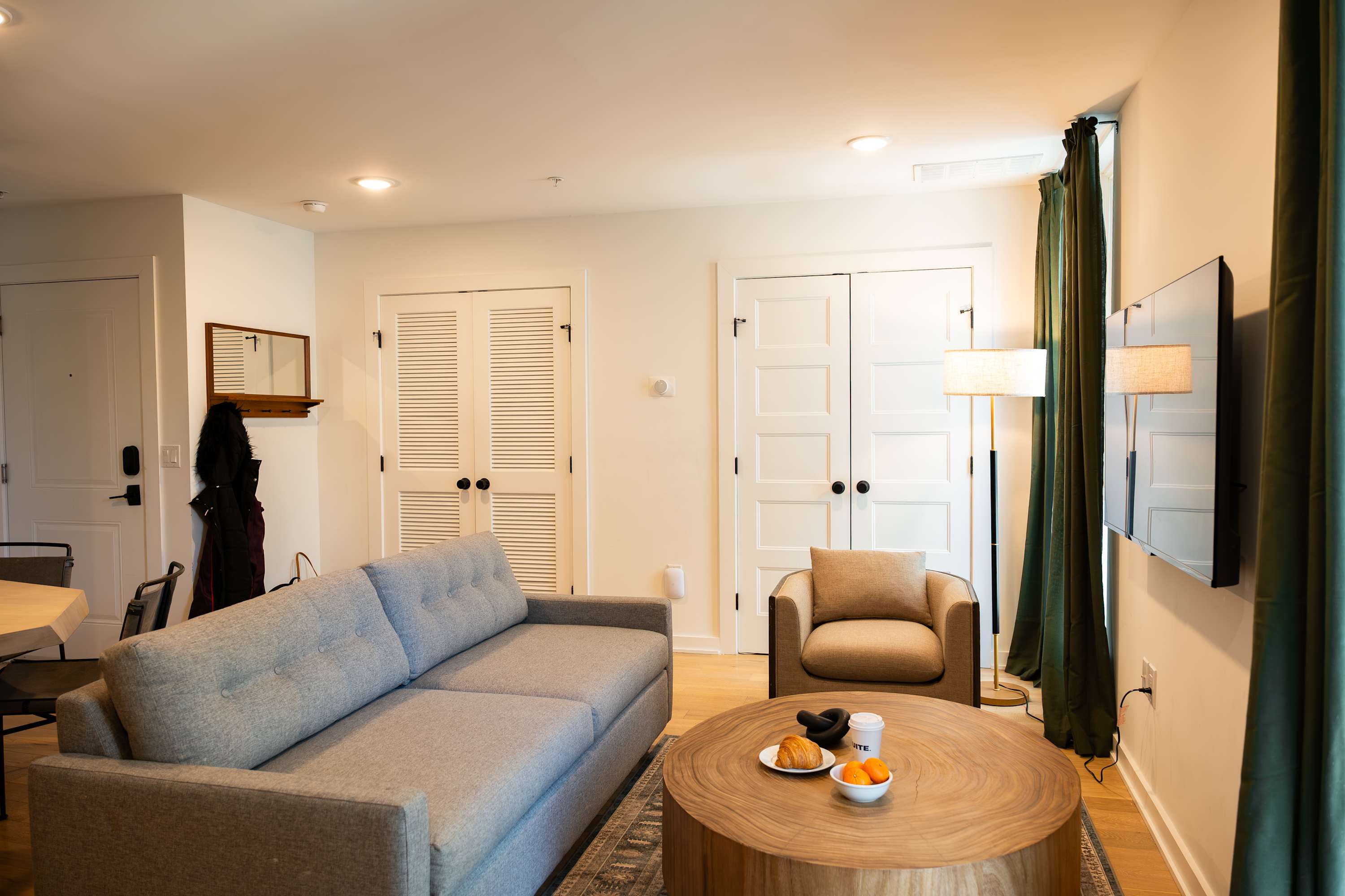 A cozy living room furnished with a gray sofa, a round wooden coffee table, and a beige armchair, illuminated by a floor lamp and featuring white doors and green curtains.