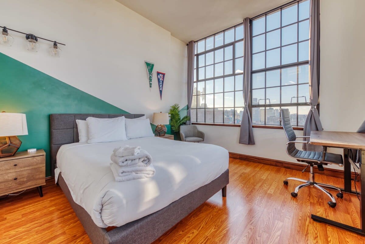 Spacious bedroom featuring a queen-sized bed with white linens, a wooden nightstand, and a dedicated workspace with a desk and office chair, all illuminated by large industrial windows.