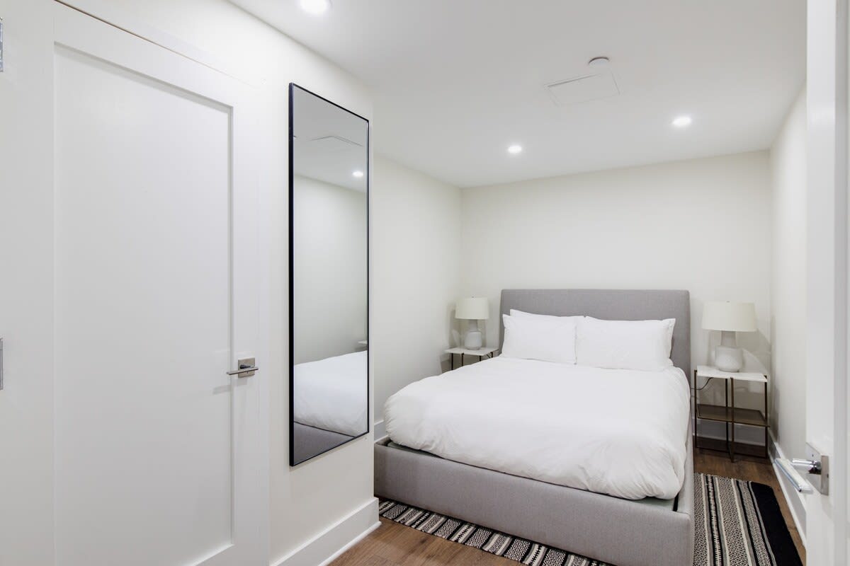 Cozy bedroom featuring a queen-sized bed with a grey upholstered headboard, white bedding, and two white nightstands with lamps, complemented by a full-length mirror on the wall.