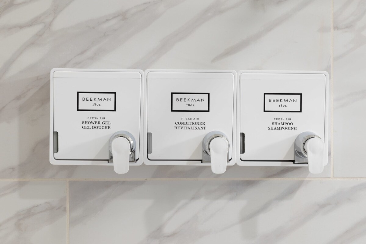 A close-up view shows three wall-mounted white dispensers labeled 'BEEKMAN 1802' for shower gel, conditioner, and shampoo, neatly installed against a white marble-patterned tiled shower wall.