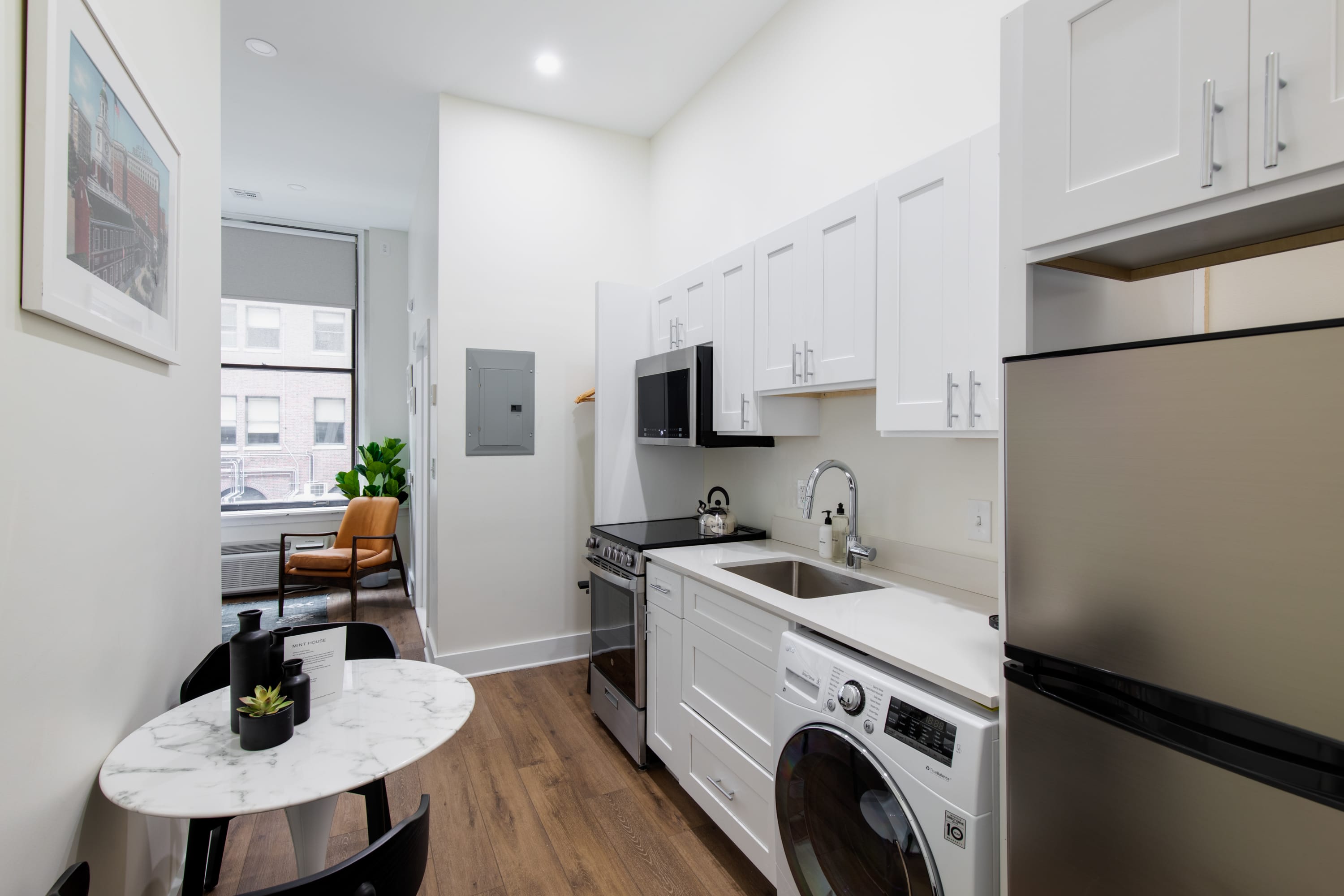 Compact modern kitchen with white shaker cabinets, stainless steel appliances including a stove, microwave, and refrigerator, and a white countertop with an integrated sink. A small round white dining table with black chairs is in the foreground.