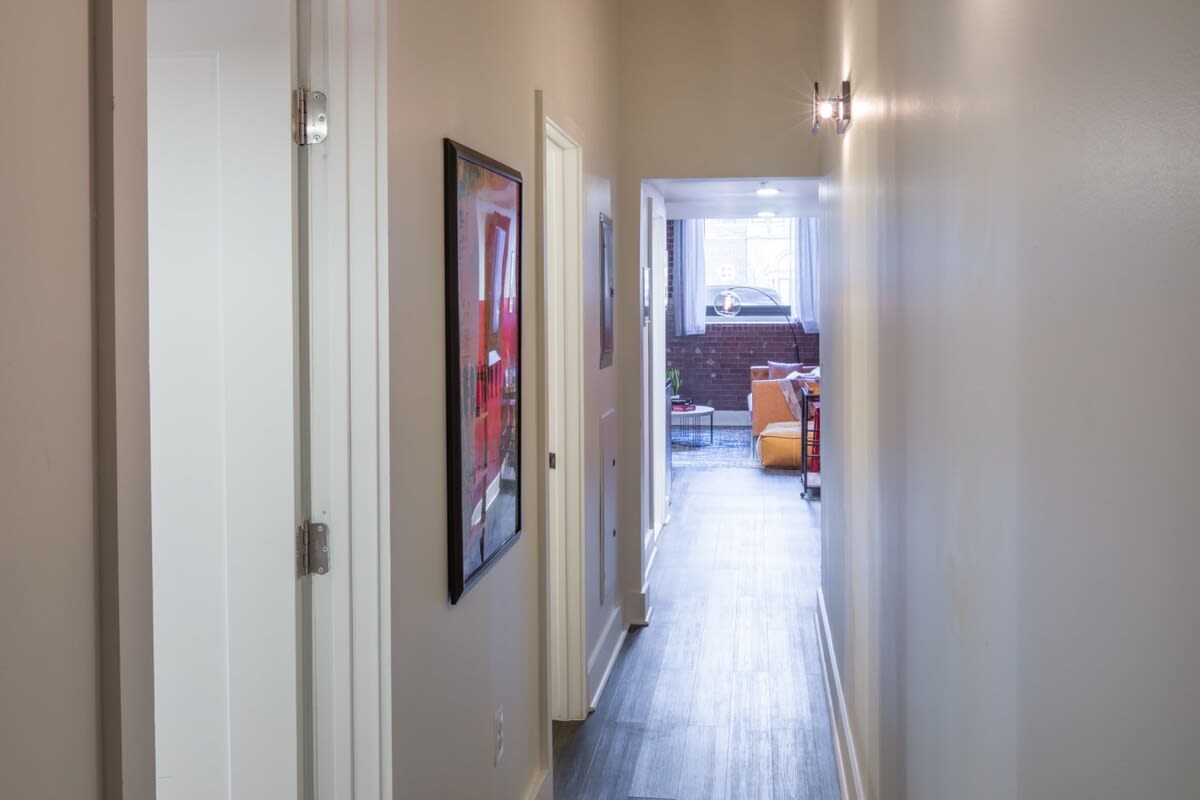 Narrow hallway with light gray walls and dark hardwood floors, leading towards the main living area visible at the far end. A framed artwork hangs on the left wall.
