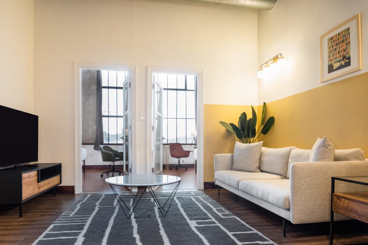 A contemporary living room with a light gray sofa, a geometric patterned rug, and a round marble-top coffee table. A black TV stand with a TV is on the left, and two open doorways lead to separate rooms with large windows.