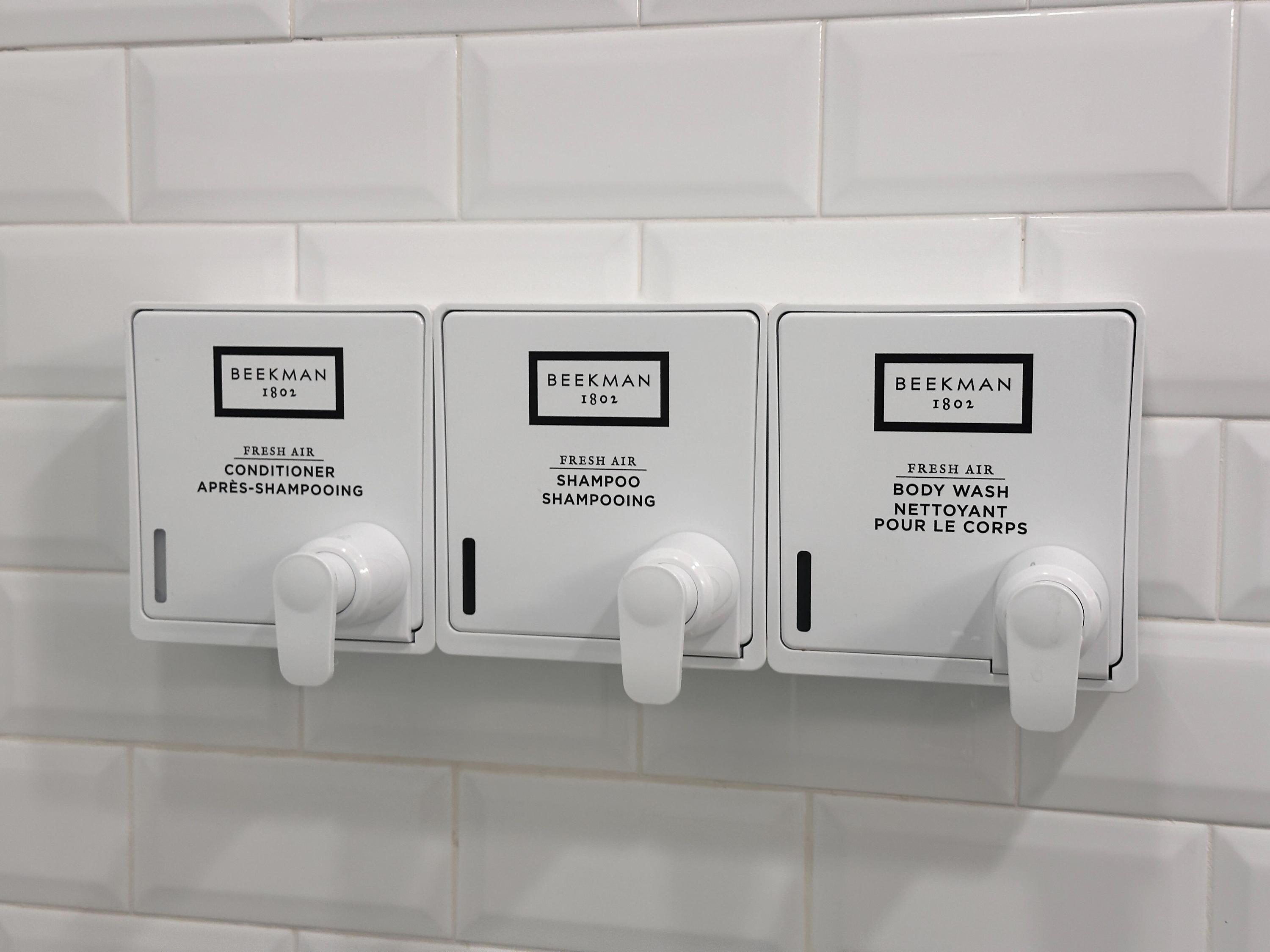 A close-up view of three wall-mounted dispensers for conditioner, shampoo, and body wash from the 'BEEKMAN 1802 FRESH AIR' line, set against a clean white subway tile wall.