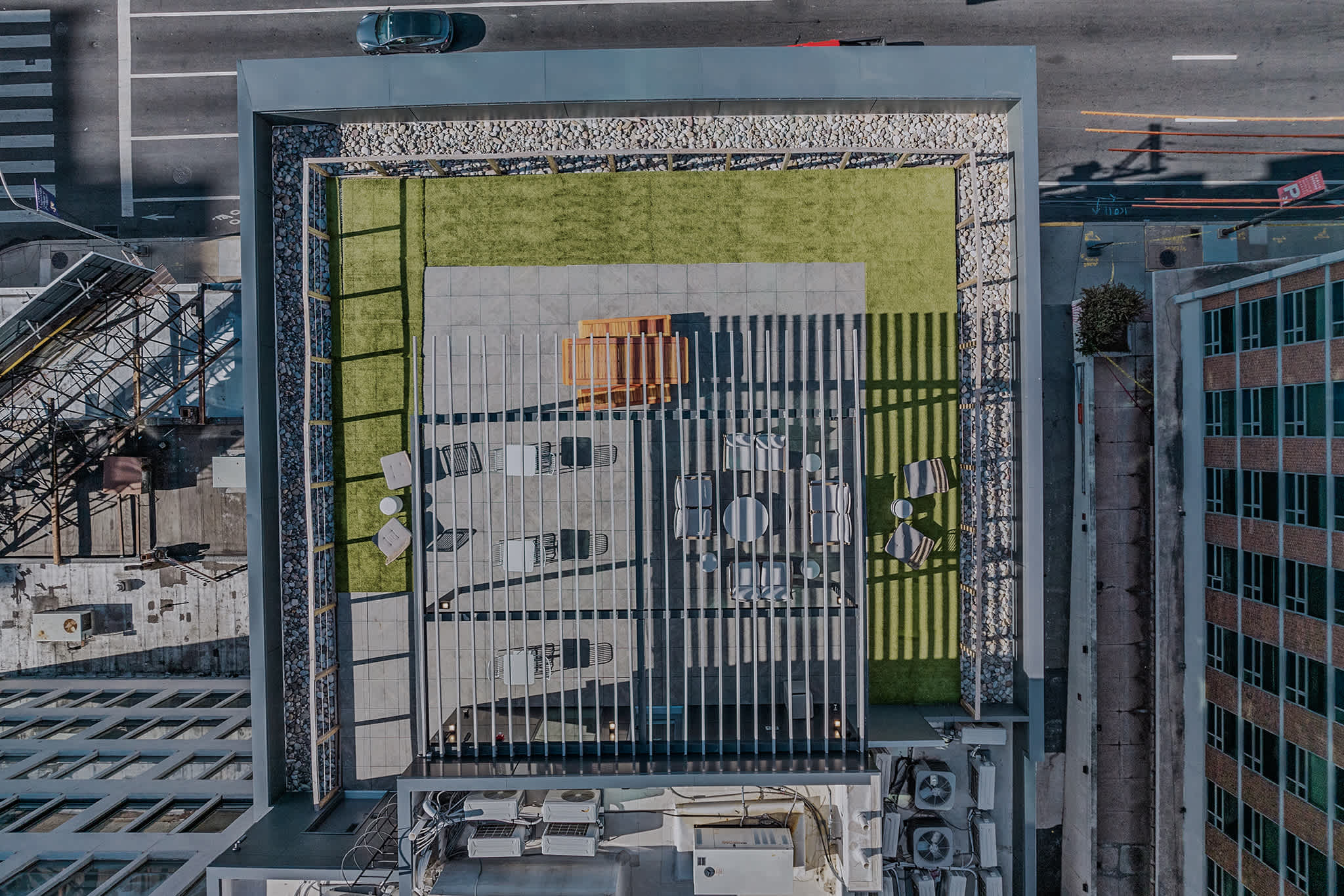 Aerial view of a modern rooftop deck featuring a large artificial turf area, concrete paving, and various outdoor seating arrangements with sun loungers and chairs, surrounded by gravel and city buildings.