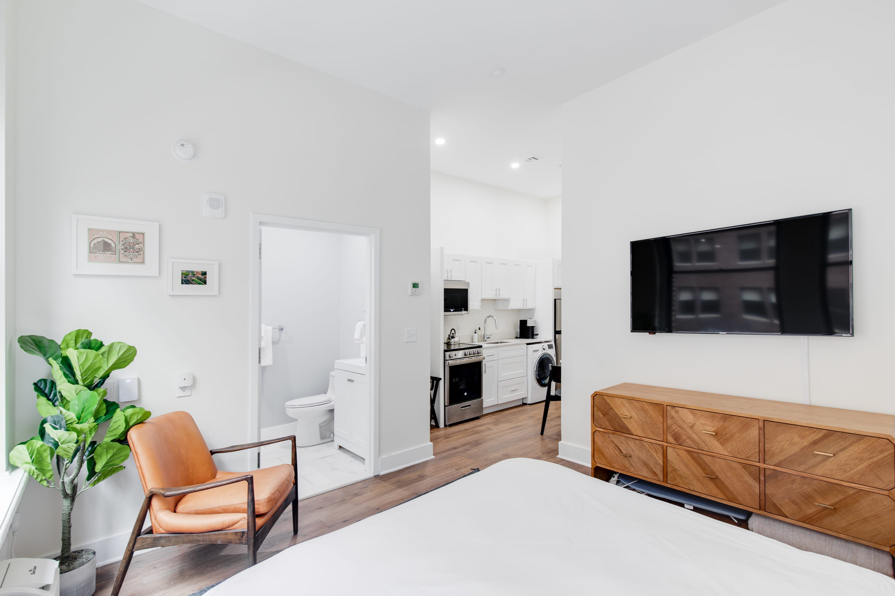 View from the bed in a bright studio apartment, showing a wall-mounted TV above a wooden dresser, an open doorway leading to a bathroom with a toilet, and a glimpse of the kitchen beyond. An orange armchair and a potted plant are in the foreground.
