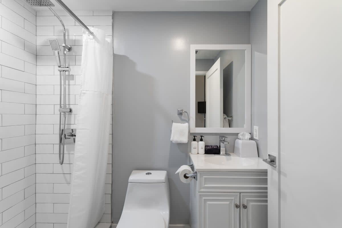 A clean, modern bathroom with light gray walls, a shower featuring white subway tile and a rain showerhead, and a white vanity with a sink and mirror. A toilet is situated next to the shower, with towels and shampoo bottles visible.