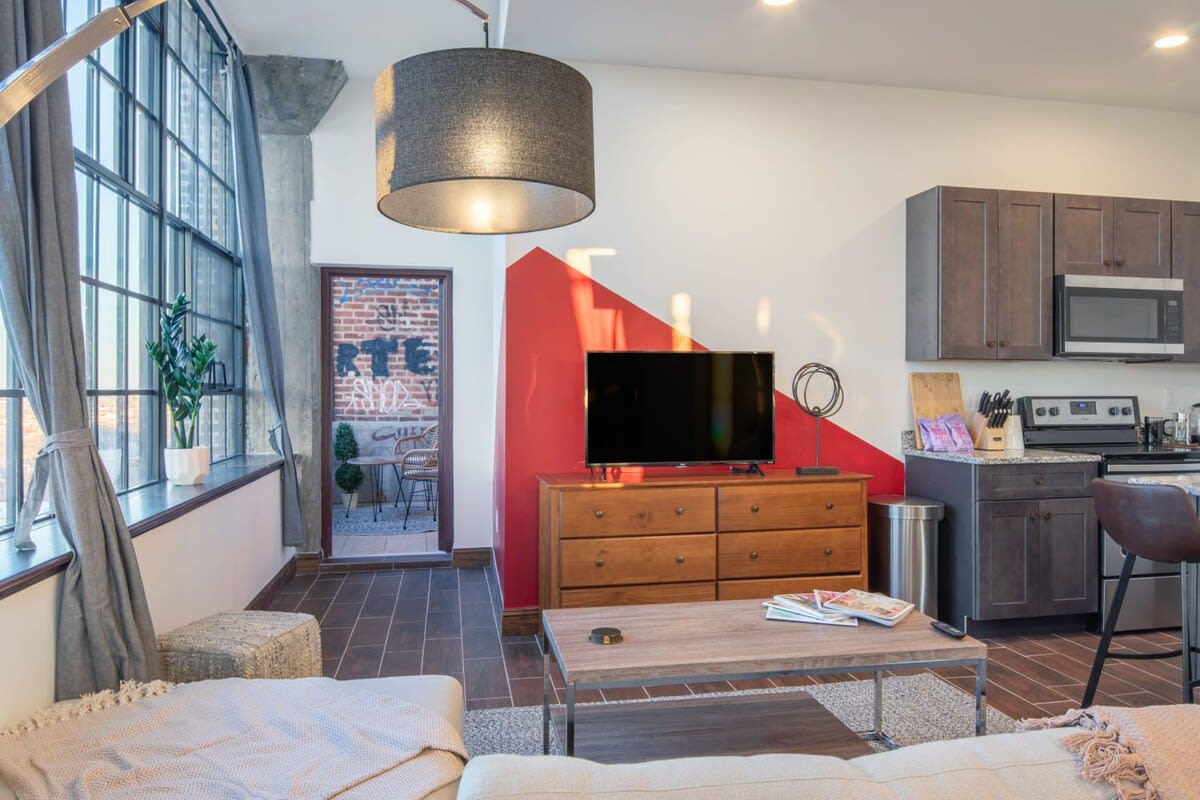Living room area with a flat-screen TV on a wooden dresser, set against a white wall with a striking red geometric accent, and an open doorway leading to a brick balcony.