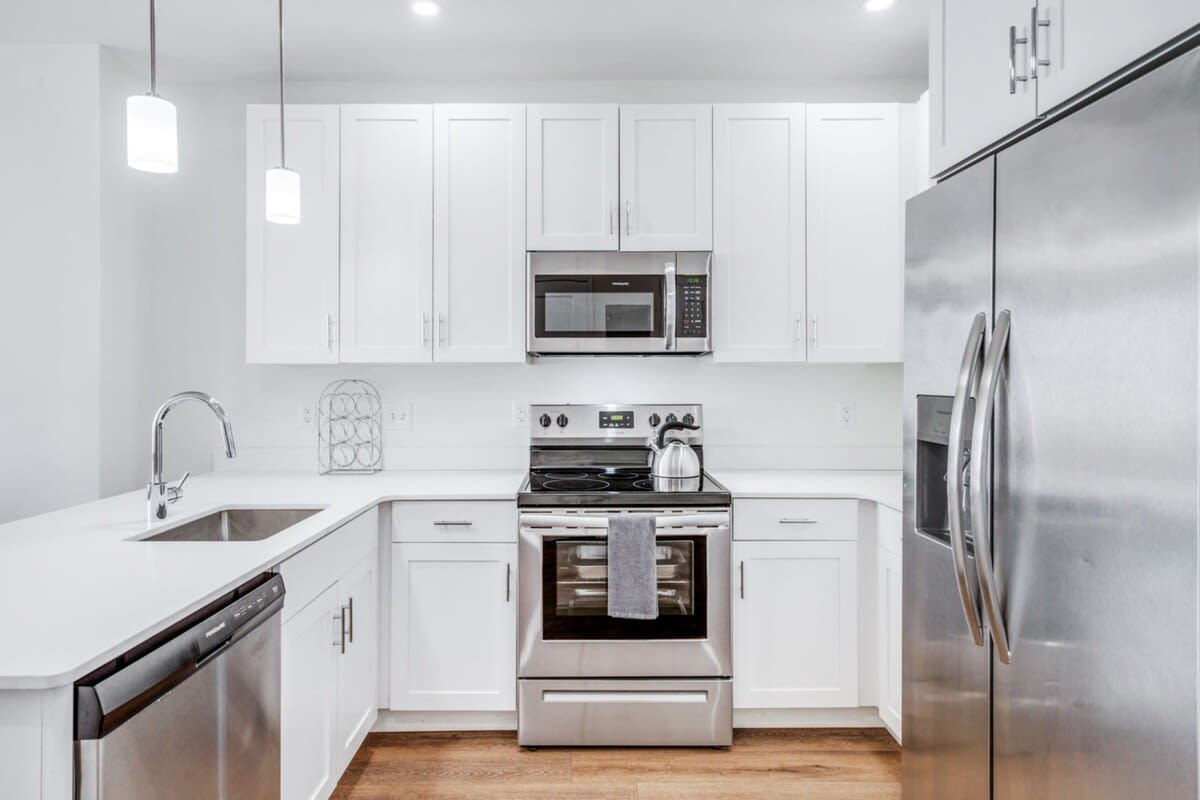 Modern kitchen with white shaker-style cabinets, white quartz countertops, and stainless steel appliances including a refrigerator, microwave, oven, and dishwasher. Pendant lights hang over the sink area.