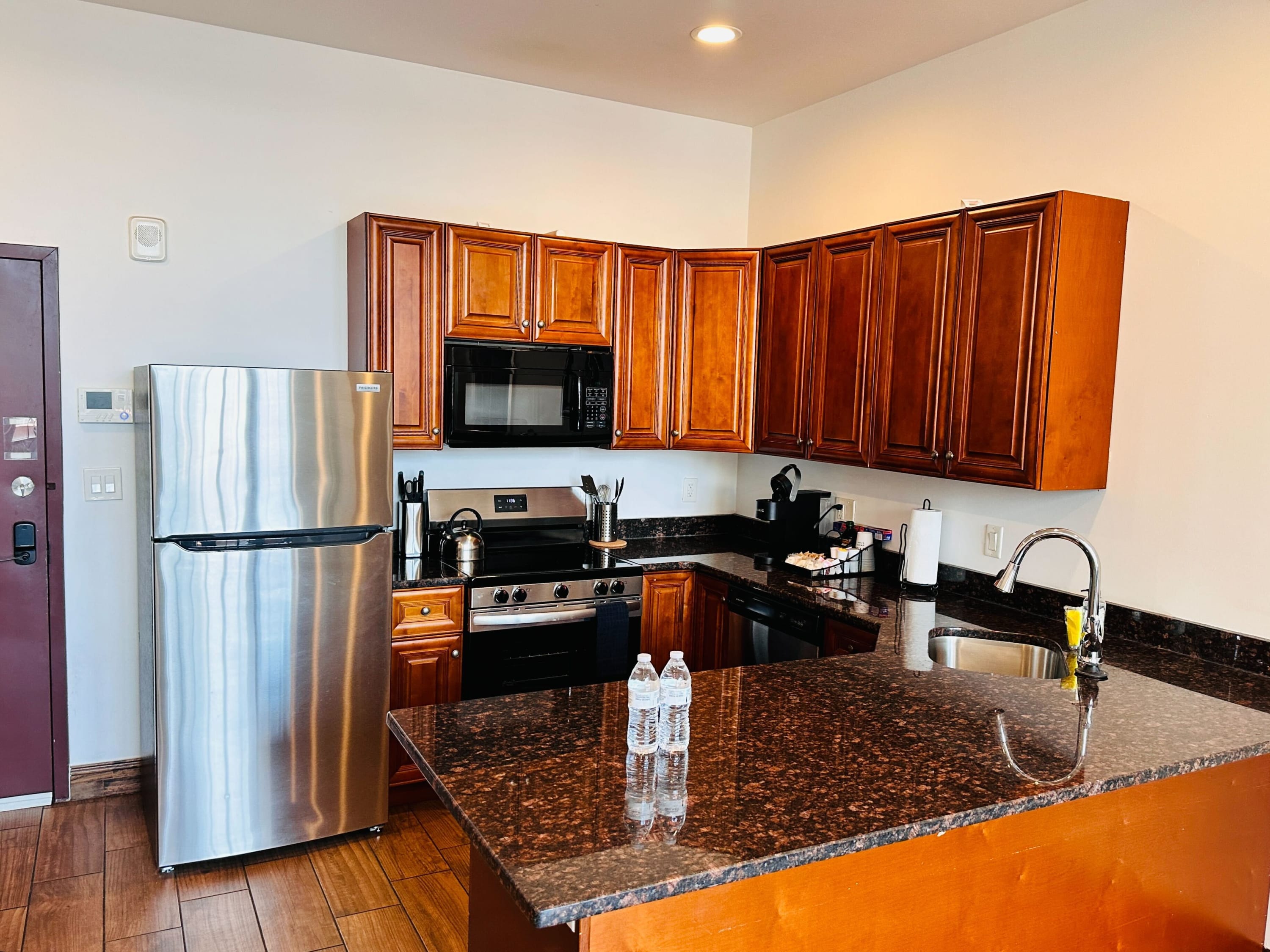 Modern kitchen with dark wood cabinetry, dark granite countertops, and stainless steel appliances including a refrigerator, microwave, and range, with a large island in the foreground.