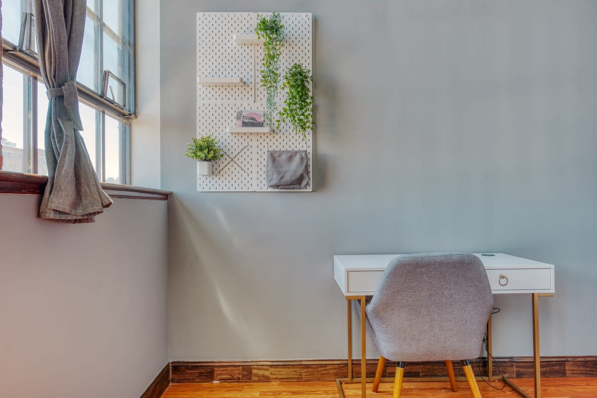 A minimalist white desk with gold accents and a gray upholstered chair sits next to a large industrial window, with a white pegboard decorated with plants on the wall above.