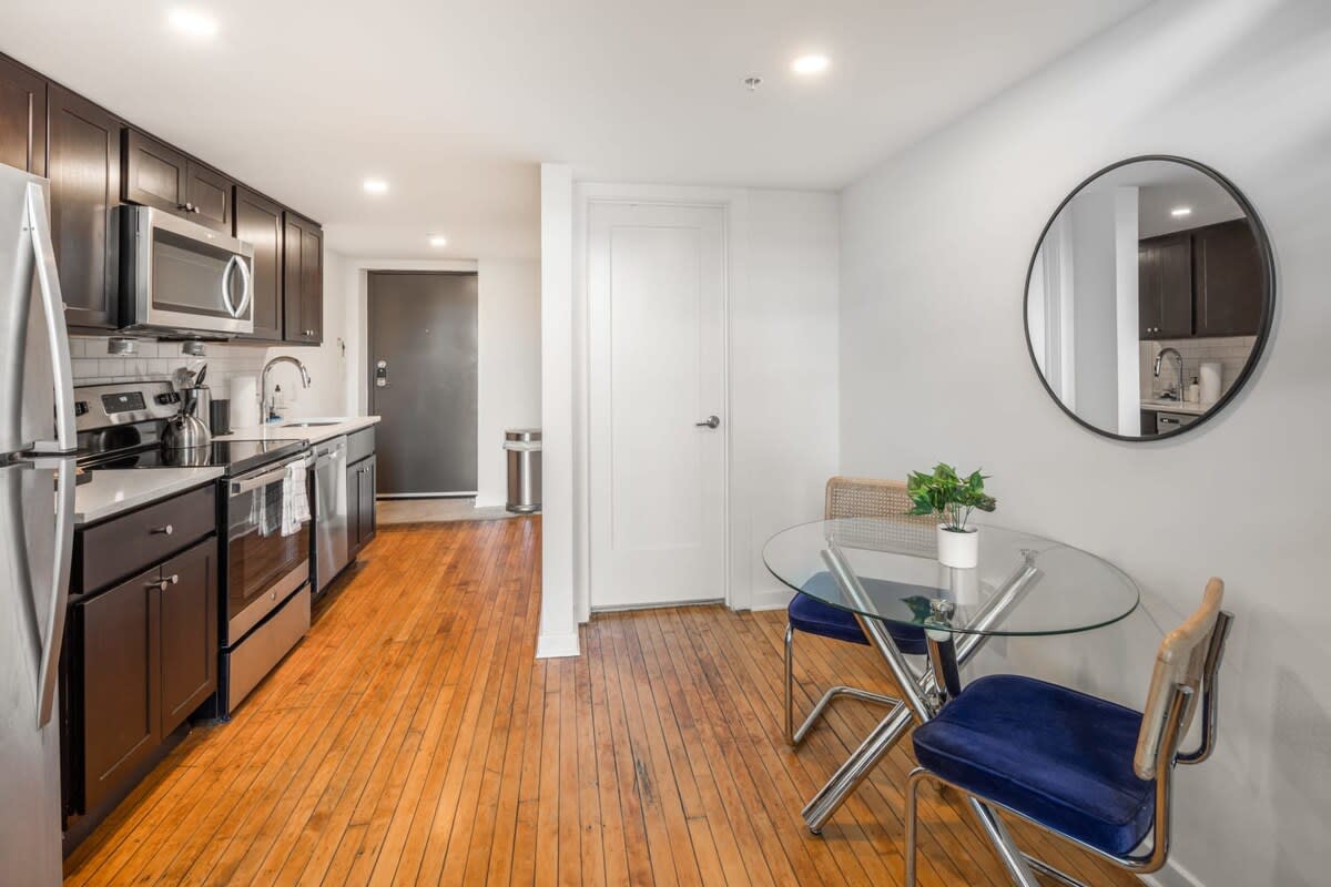 An open-concept space showcasing a modern kitchen with dark cabinetry and stainless steel appliances, flowing into a dining area with a glass-top table and two chairs, all on warm hardwood floors.