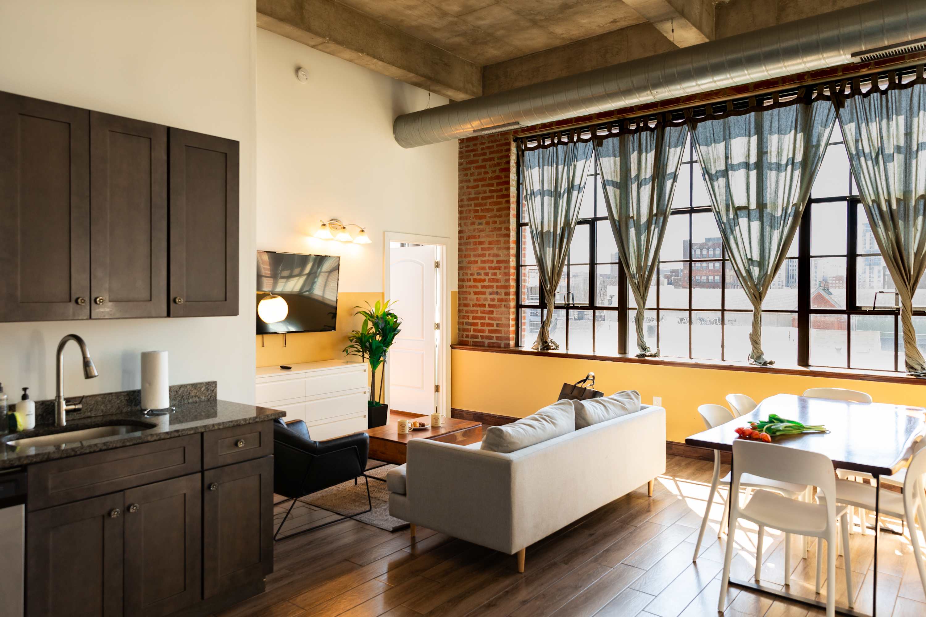 An open-concept living area with a modern kitchen, light sofa, TV, and dining table. Large industrial windows with sheer curtains overlook the city, complemented by exposed brick and a concrete ceiling, all on hardwood floors.