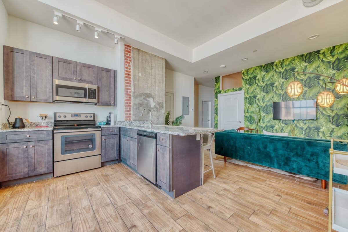 A spacious kitchen featuring dark wood cabinets, stainless steel appliances, granite countertops, and a large island with bar seating, seamlessly connecting to the living area with a green accent wall.