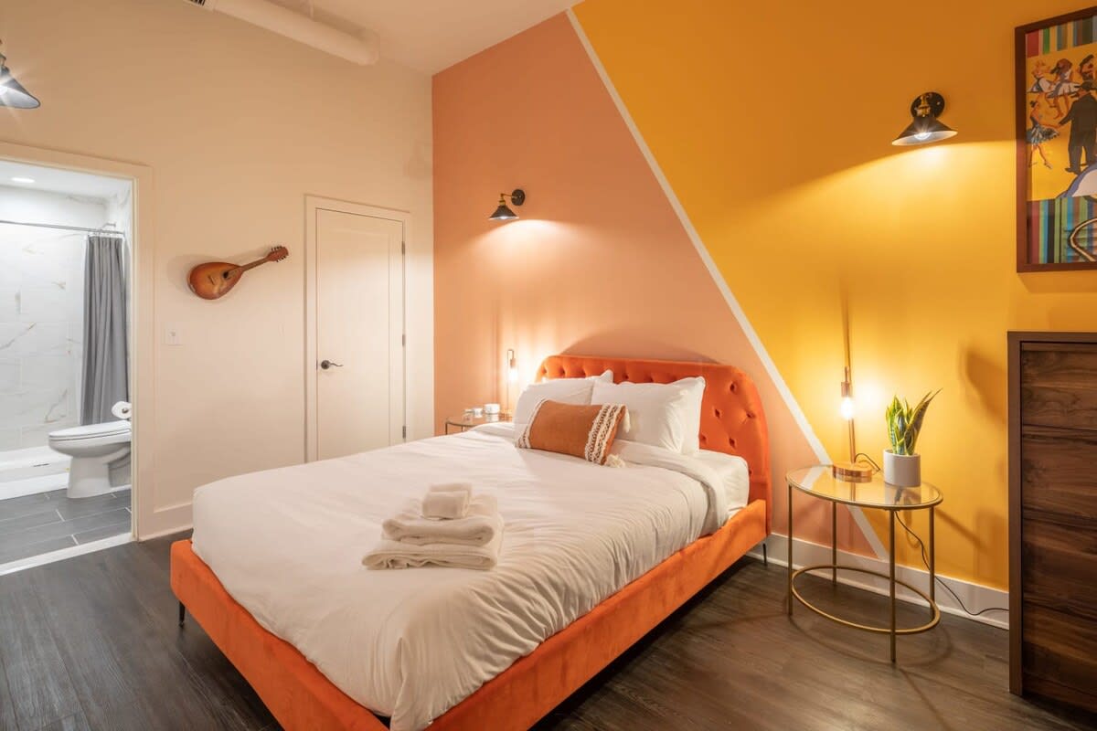 Vibrant bedroom featuring an orange upholstered bed with white linens and a patterned throw pillow. The walls are painted with a geometric design in peach and yellow, complemented by a dark wood dresser and a gold-framed side table with a plant. A bathroom entrance is visible to the left.
