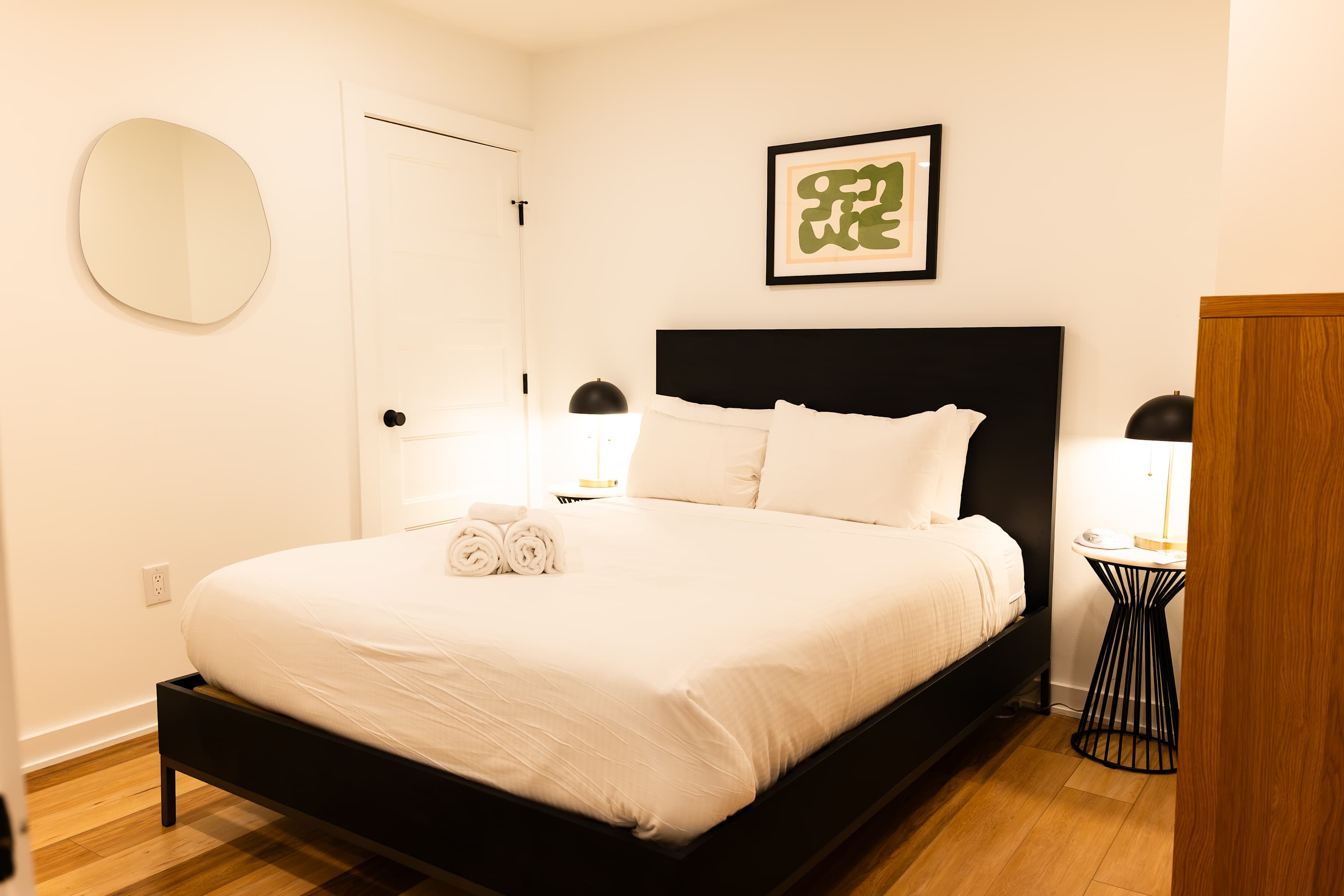A bright bedroom with a queen-sized bed, white linens, a black headboard, and light-colored walls, featuring a round mirror and a framed abstract artwork above the bed.