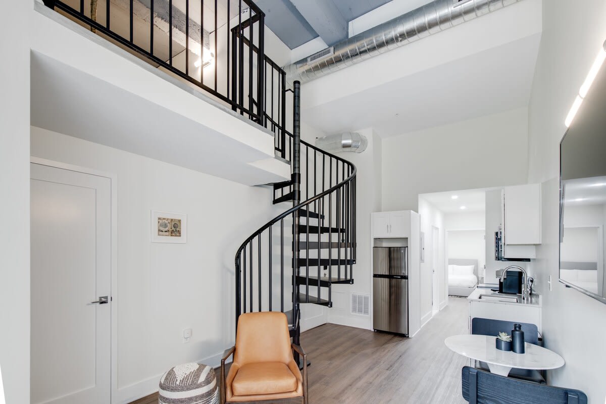 Open-concept living space featuring a black spiral staircase, a comfortable brown leather armchair, and a view into the kitchen and a hallway leading to a bedroom.