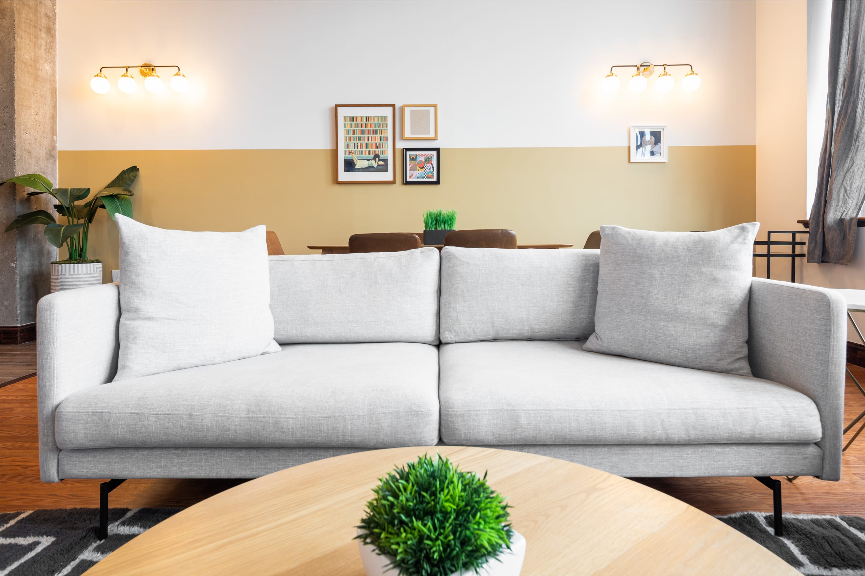 Close-up view of a light gray sofa with white throw pillows, facing a round wooden coffee table with a small green plant. The background shows a dining area with framed artwork on a two-tone wall.