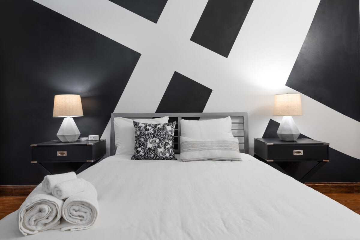 Modern bedroom with a bed flanked by two black nightstands and matching table lamps, set against a bold black and white geometric accent wall. White bedding and decorative pillows complete the contemporary look.