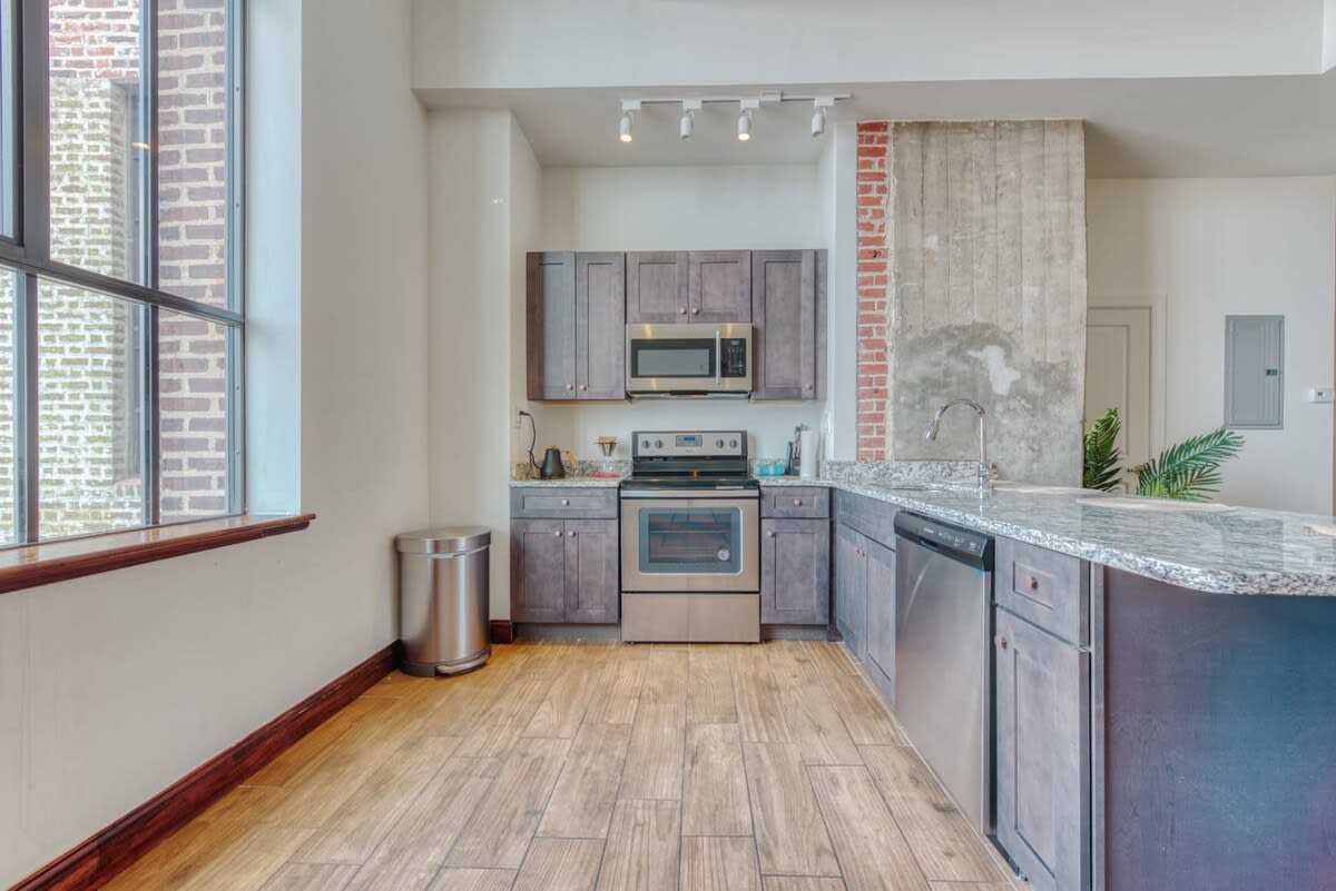 A modern kitchen with dark wood cabinetry, stainless steel appliances including a microwave, oven, and dishwasher, granite countertops, and a large industrial-style window with exposed brick.