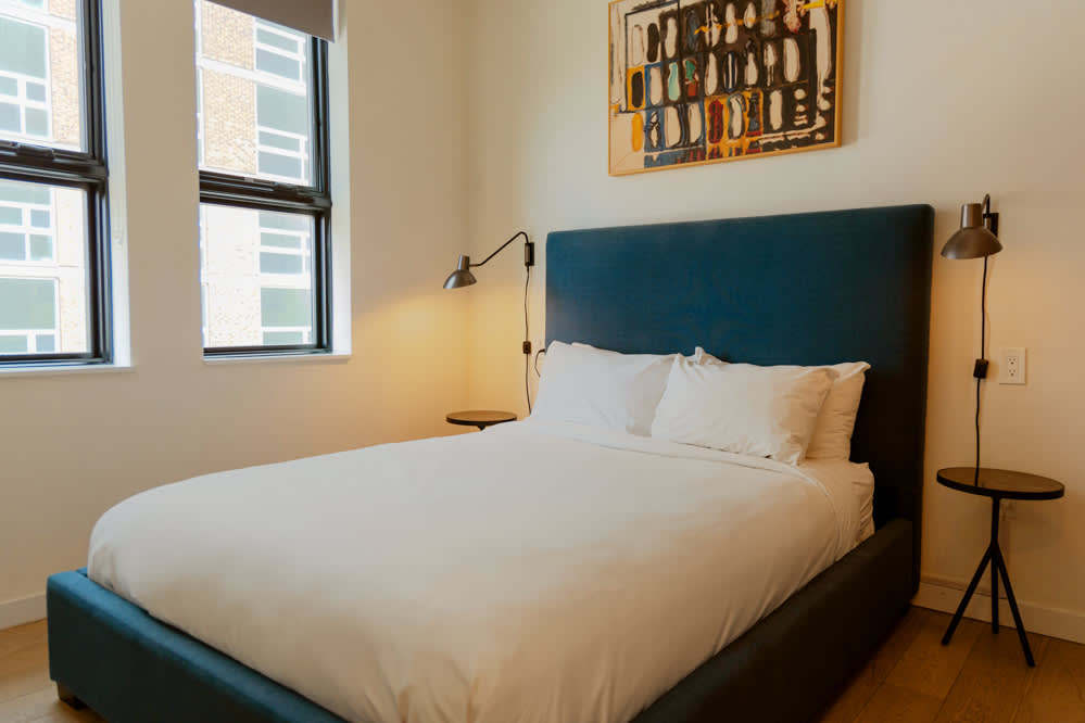 A cozy bedroom features a queen-sized bed with a dark blue upholstered headboard and white bedding, flanked by two small round black nightstands with wall-mounted sconces. A colorful abstract painting hangs above the bed, and two large windows provide natural light.