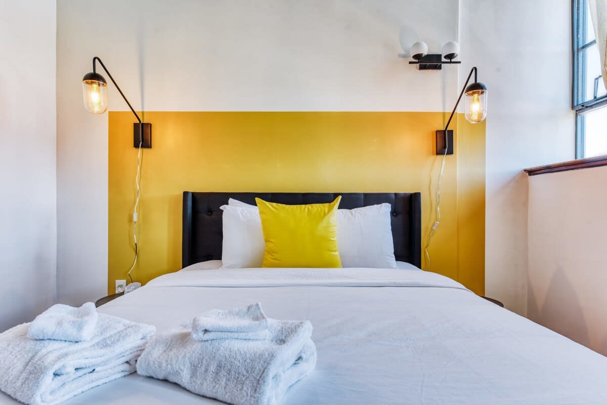 A bright bedroom with a black upholstered headboard against a striking yellow accent wall, flanked by modern black wall sconces with exposed bulbs. The bed is dressed in crisp white linens with a single yellow throw pillow, and white folded towels are neatly placed at the foot of the bed.