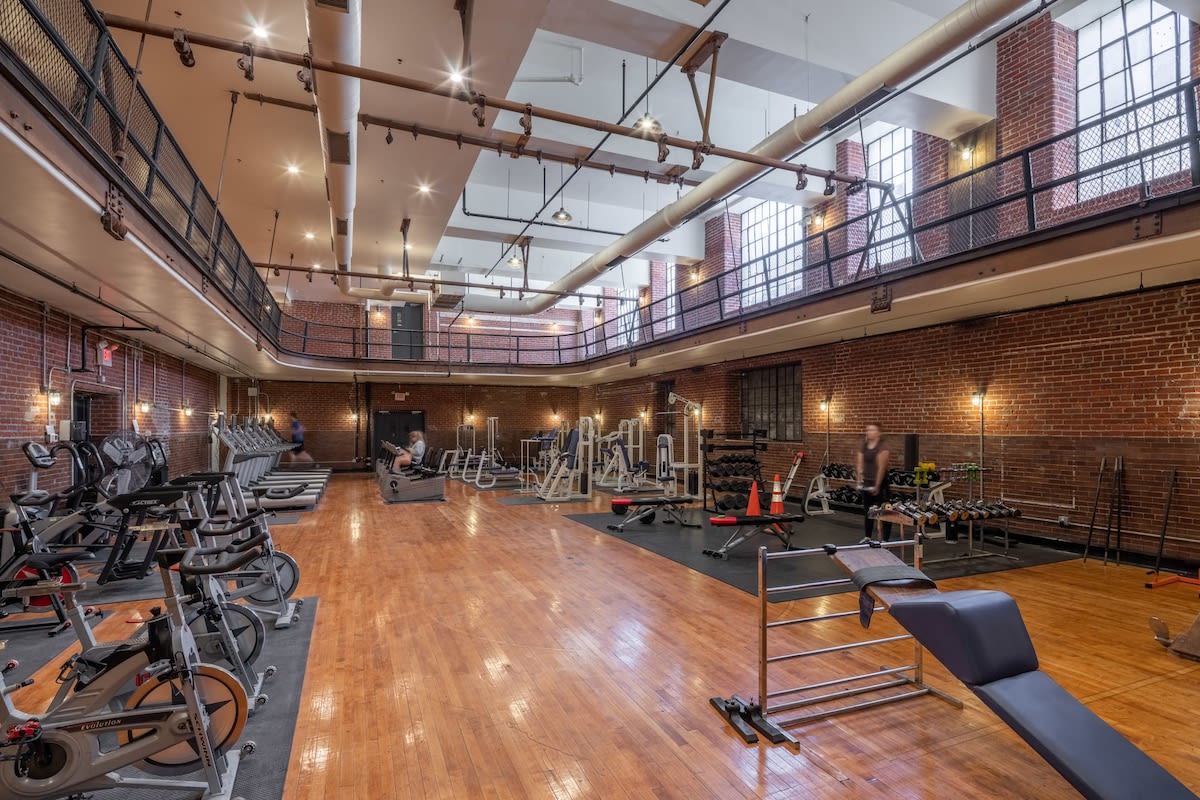 A spacious, industrial-style gym with high ceilings, exposed brick walls, and large windows. It features various exercise equipment including treadmills, ellipticals, stationary bikes, and weight machines, all set on a polished hardwood floor.