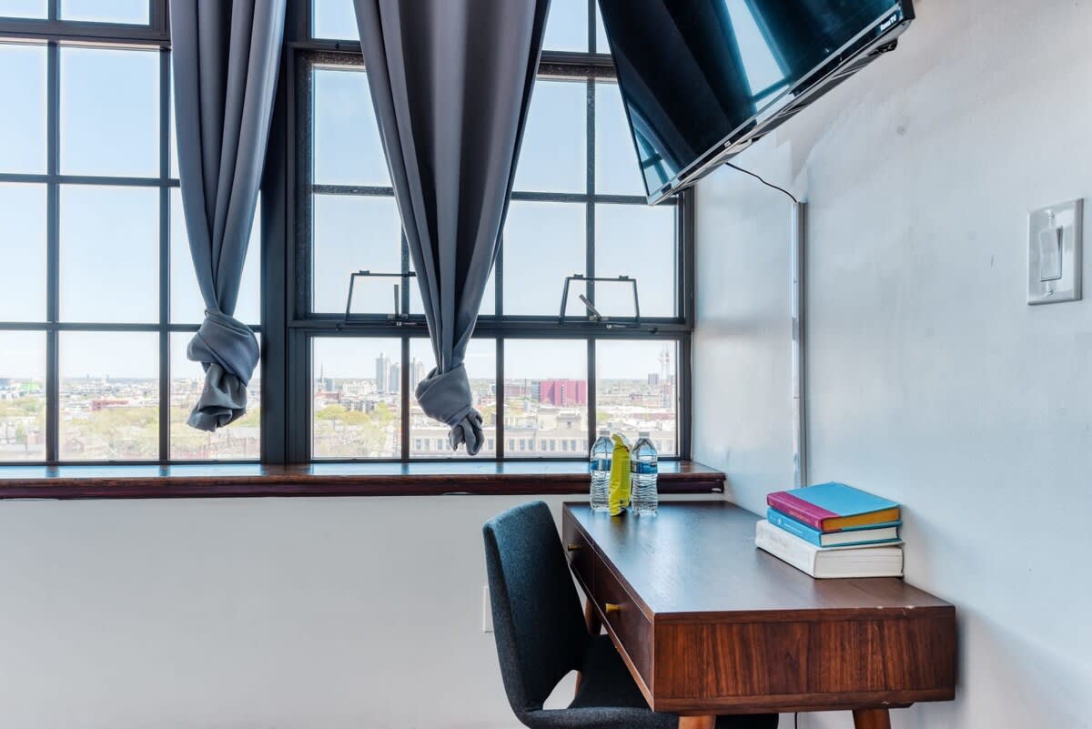 Dedicated workspace with a mid-century modern wooden desk and a dark grey chair positioned by large industrial windows. Bottles of water and books are neatly arranged on the desk, with a wall-mounted TV above.