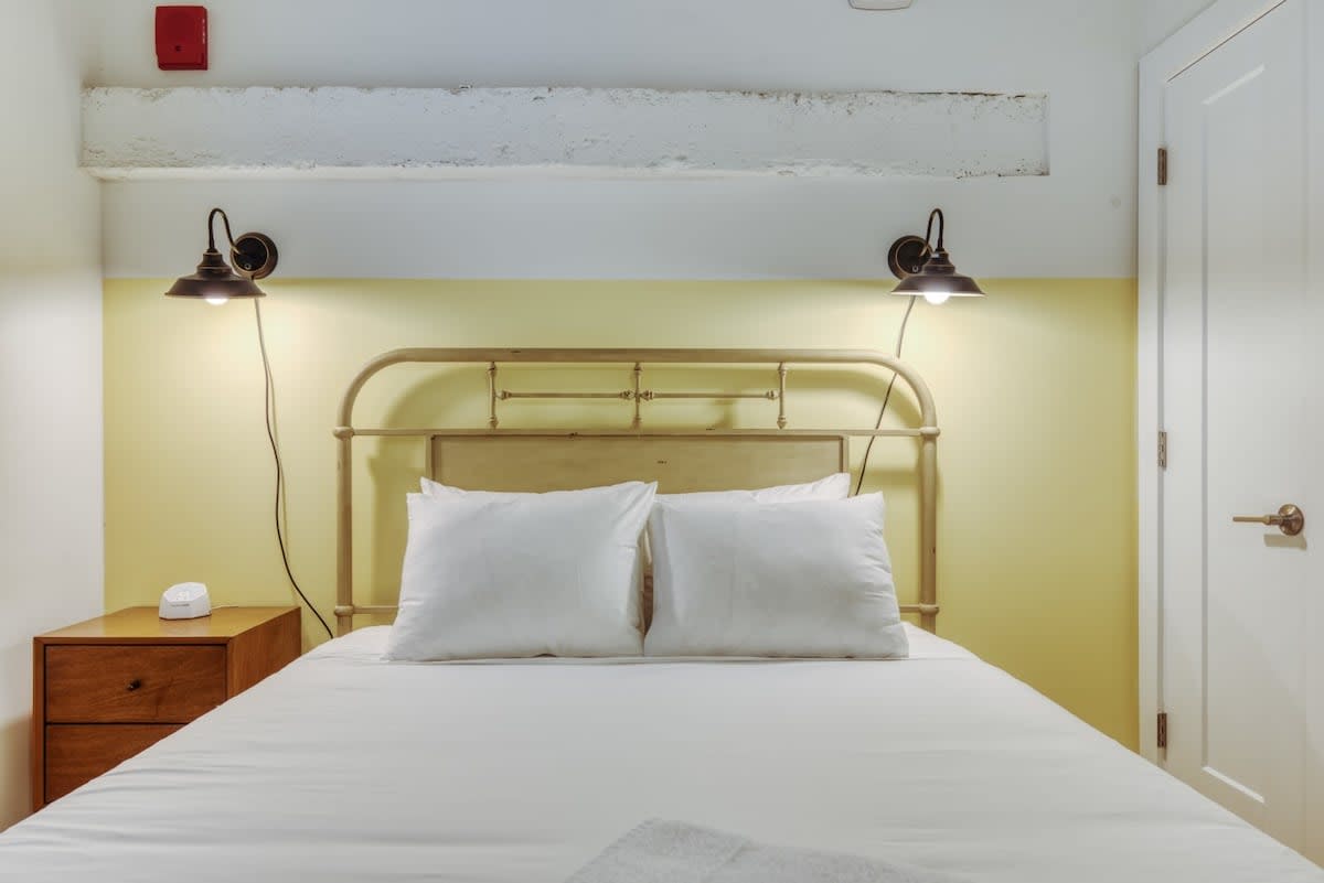 Cozy bedroom with a vintage-style metal bed frame, crisp white bedding, and two industrial-style wall sconces illuminating a light yellow accent wall. A simple wooden nightstand with a white alarm clock sits beside the bed.