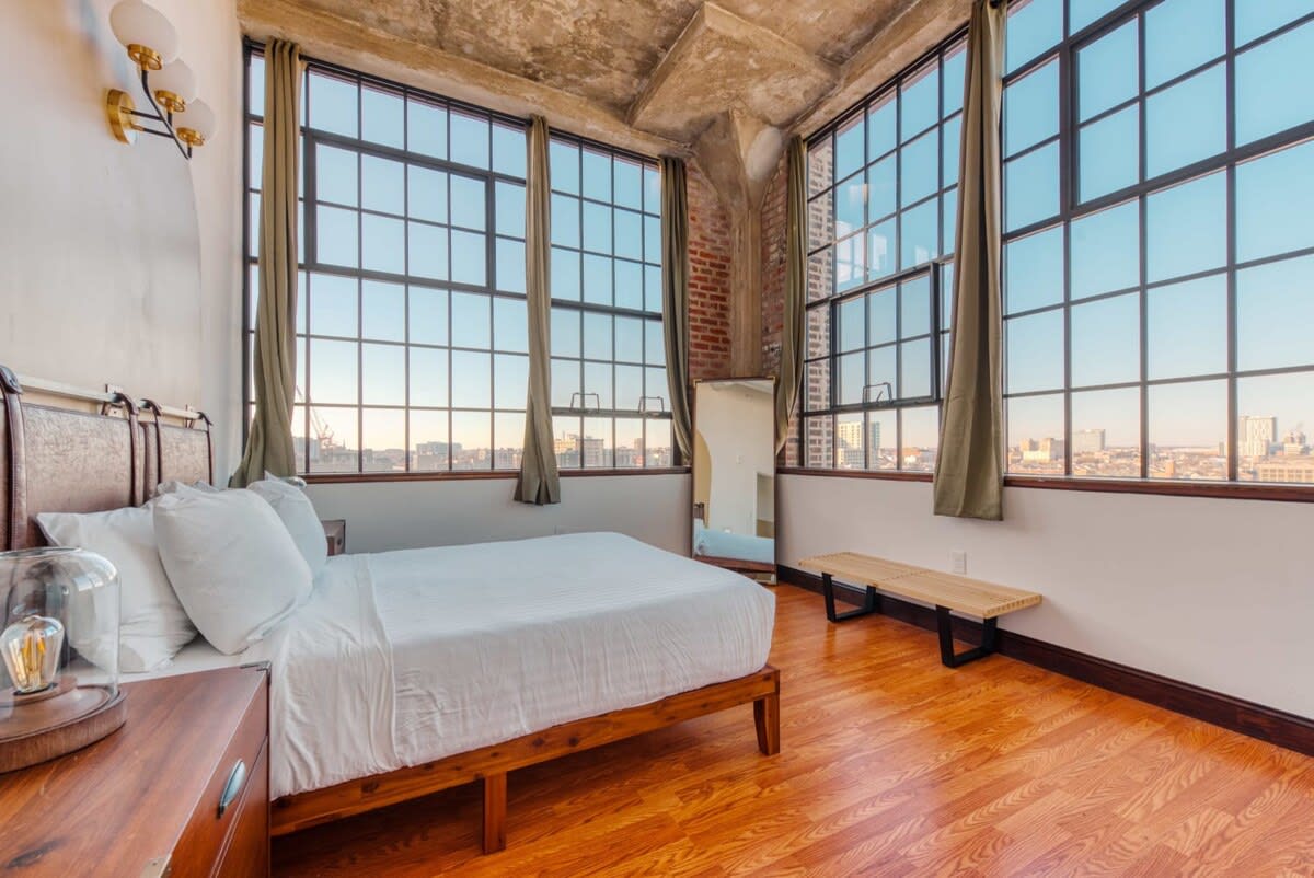 Bright bedroom with a queen-sized bed, light wood frame, and hardwood floors, featuring large industrial-style windows with city views and a concrete ceiling.