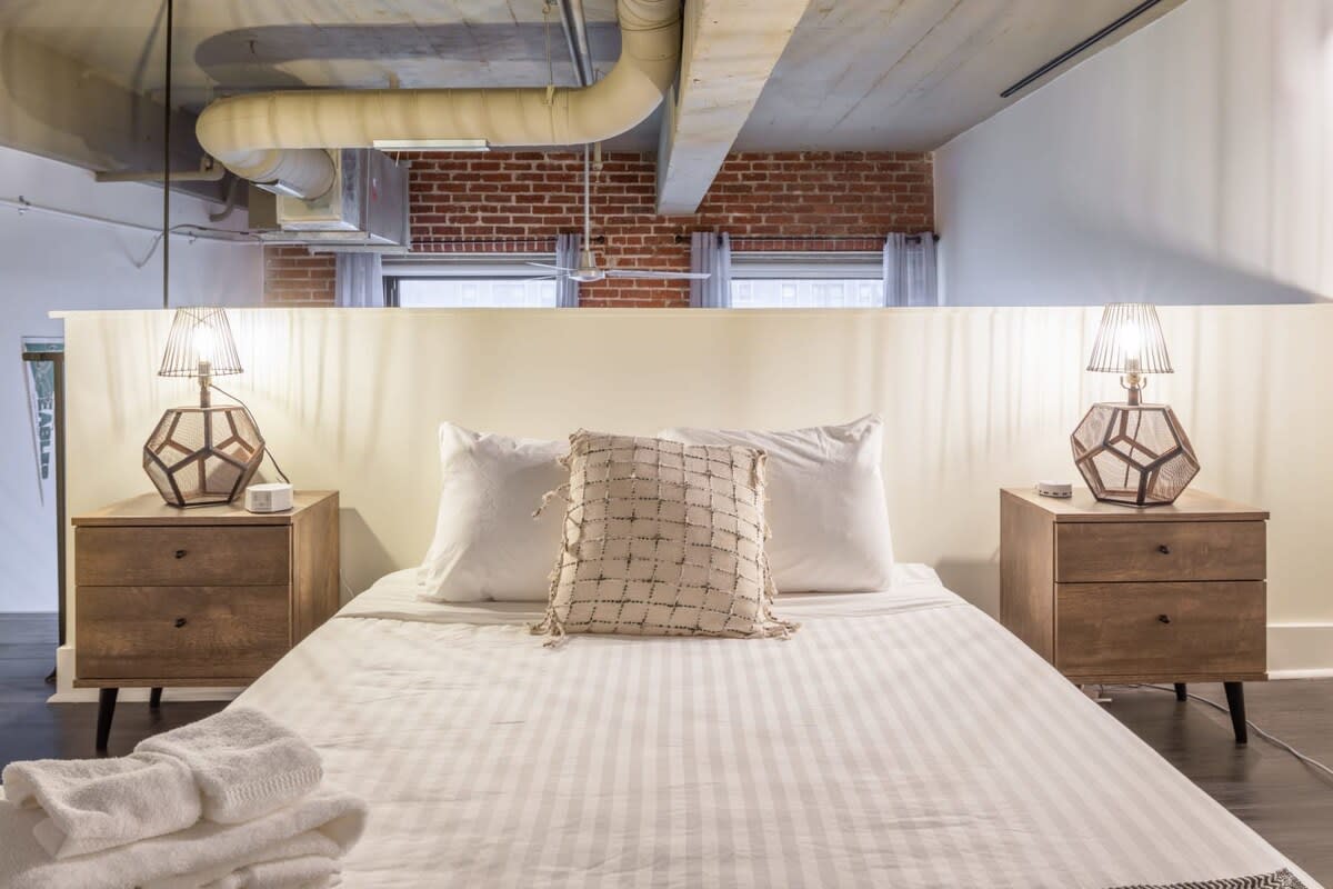 Cozy bedroom with a queen-sized bed, two wooden nightstands, and geometric lamps. A partial wall acts as a headboard, with exposed brick and industrial ductwork visible above.