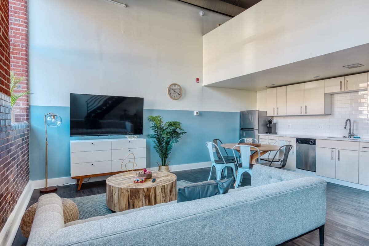 Open-concept living area and kitchen with a light blue and white two-tone wall, featuring a large flat-screen TV on a white console, a light gray sofa, and a round wooden coffee table. The kitchen has white cabinets and stainless steel appliances.