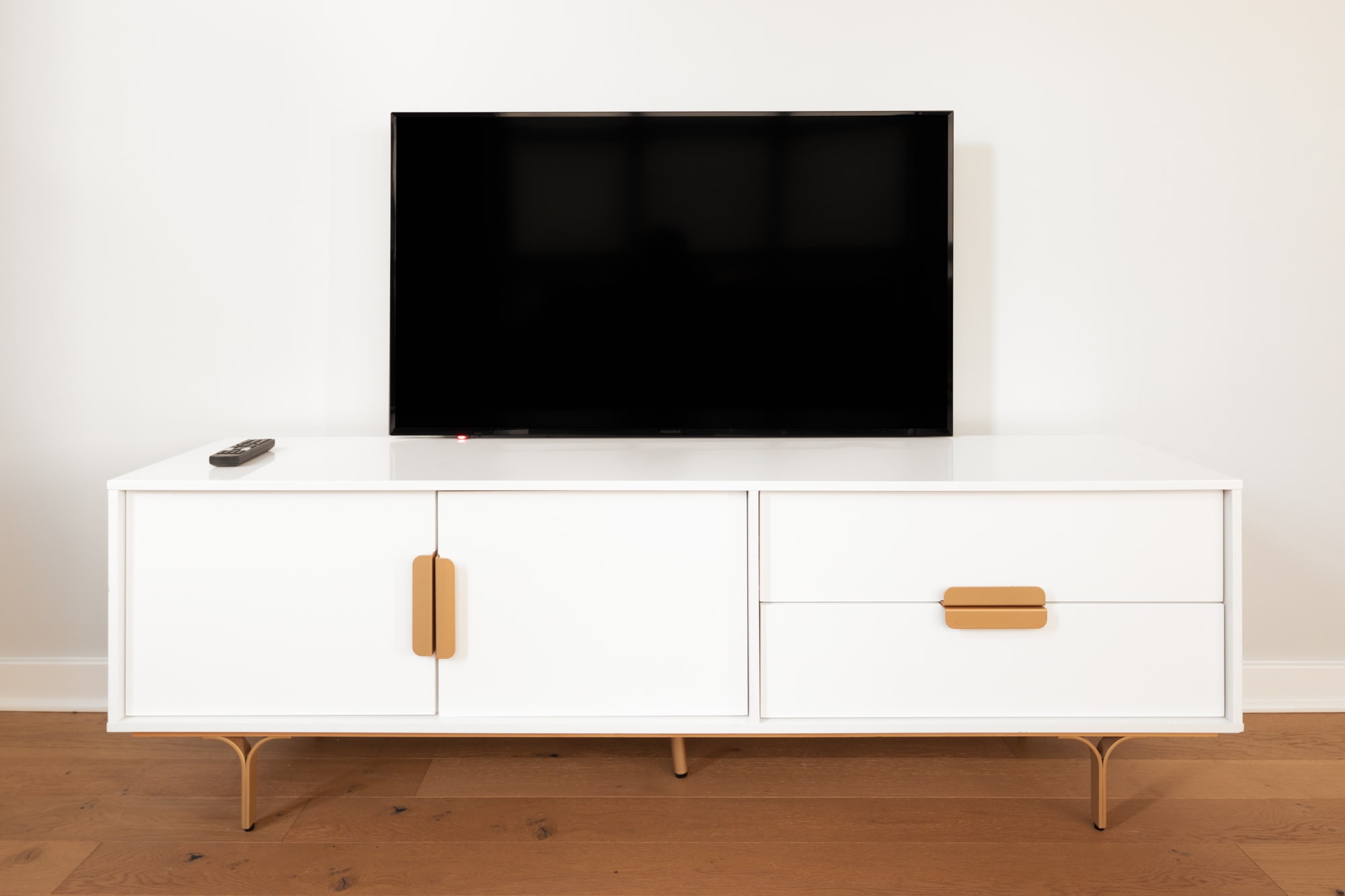 Detail shot of a black flat-screen TV resting on a white media console with gold hardware and legs. A remote control is visible on the console, set against a plain white wall.