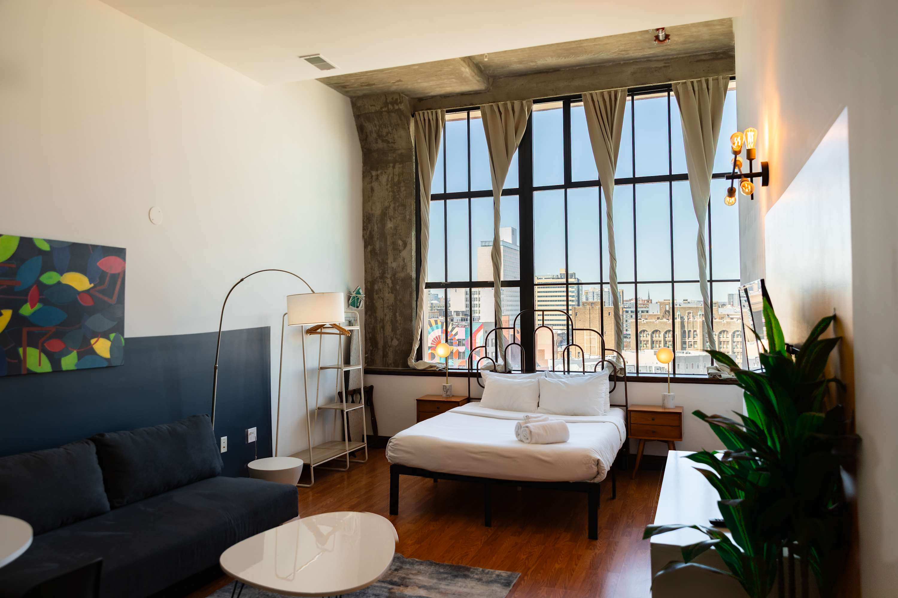 Bright studio apartment with a metal-frame queen bed, a dark gray sofa, and a floor lamp, set against a backdrop of large industrial-style windows offering city views. The room features hardwood floors and a concrete ceiling.