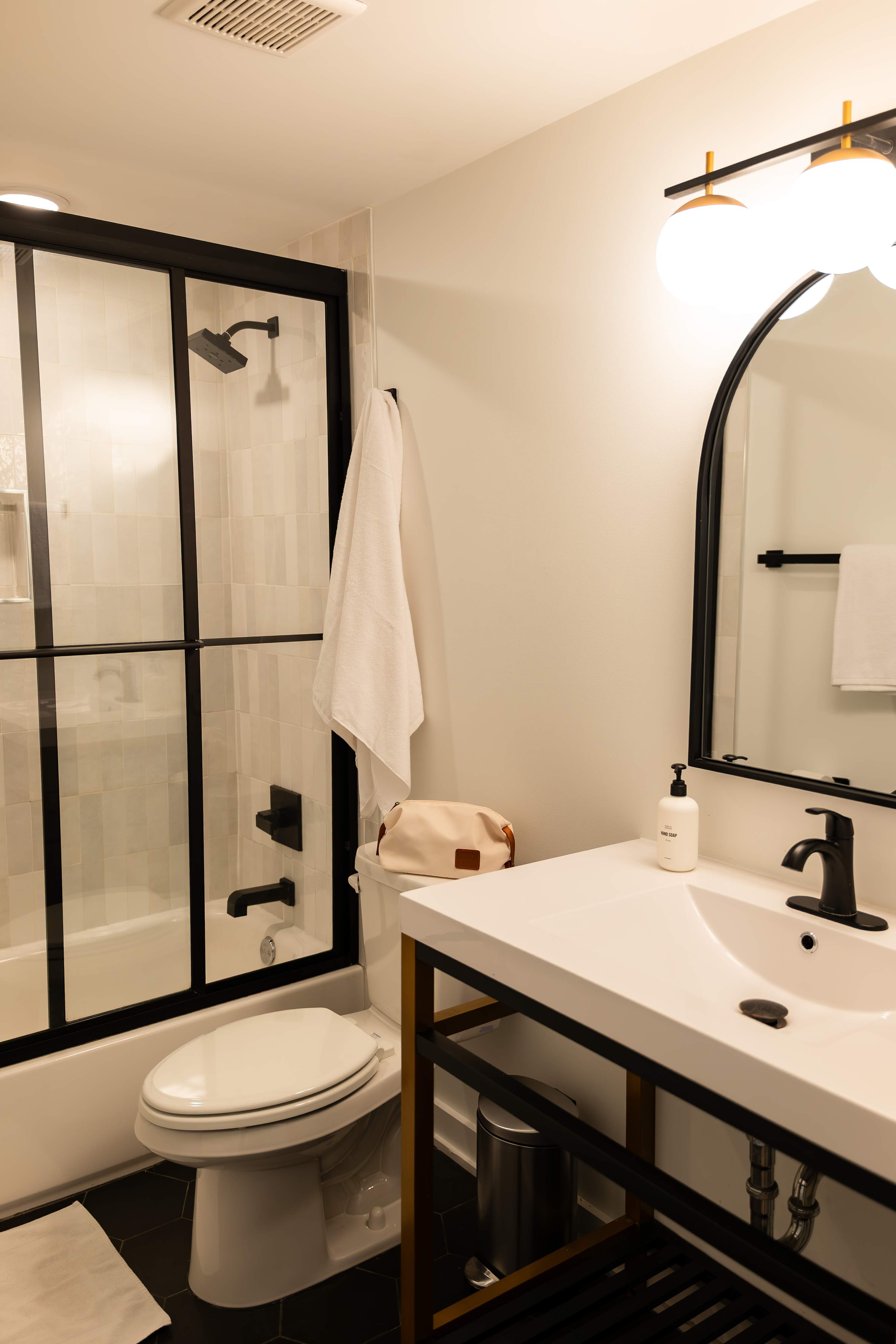 A contemporary bathroom with a black-framed glass shower enclosure, a white toilet, and a modern vanity with a black and gold frame, a white sink, and an arched mirror.