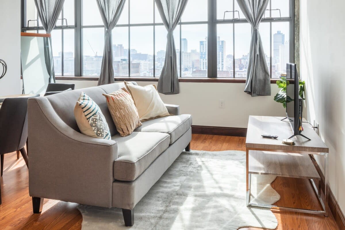 Close-up of the inviting living room, showcasing a light grey sofa with patterned throw pillows, a flat-screen TV on a sleek wooden console, and a soft white area rug over warm hardwood floors, all bathed in natural light from large windows.