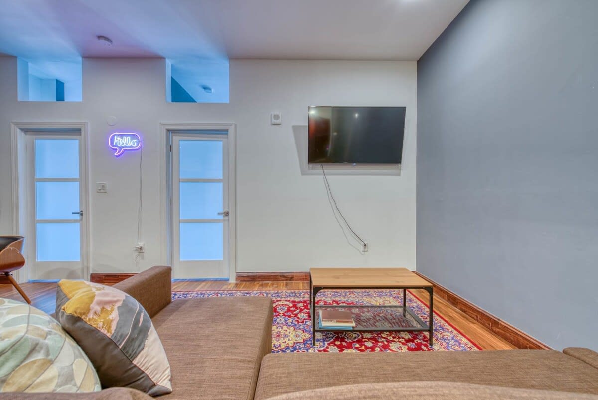 A cozy living area featuring a brown sofa with decorative pillows, a small black coffee table, and a wall-mounted flat-screen TV, set on a vibrant red patterned area rug over hardwood floors.
