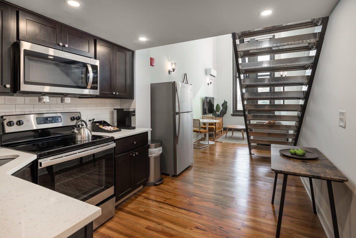 An open-concept space featuring a dark wood kitchen with stainless steel appliances, including a refrigerator, stove, microwave, and dishwasher. Hardwood floors extend into the living area, which has a sofa and a black metal staircase leading upstairs.