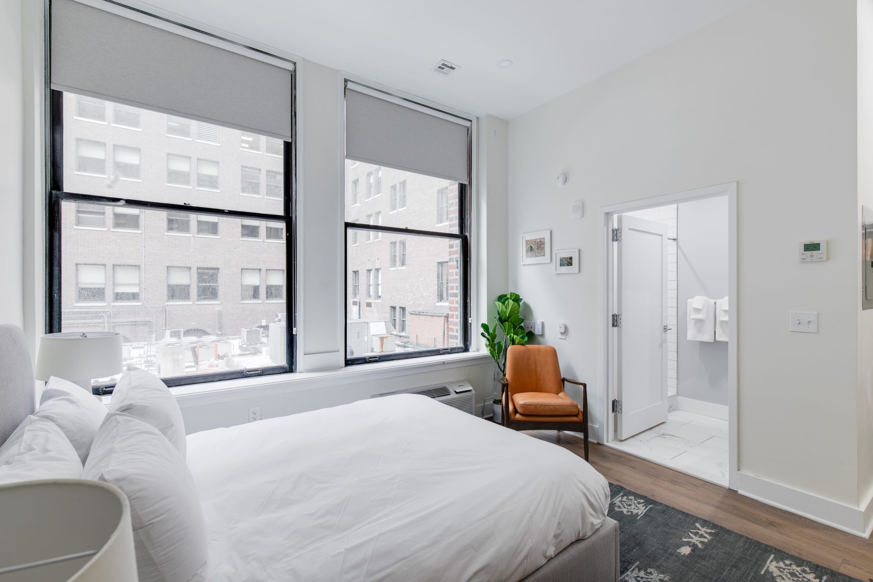 Bright bedroom featuring a gray upholstered bed with white bedding, large windows with gray blinds, and an orange armchair next to a potted plant. An open doorway reveals a white-tiled bathroom with a shower.