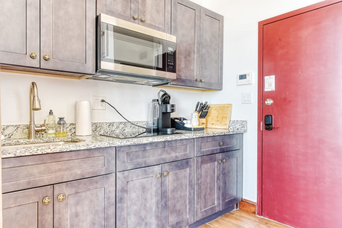 Compact kitchen area featuring grey shaker-style cabinets with ornate gold hardware, light granite countertops, and a stainless steel microwave. A coffee maker, sink, and various kitchen essentials are visible, with a red entrance door to the right.