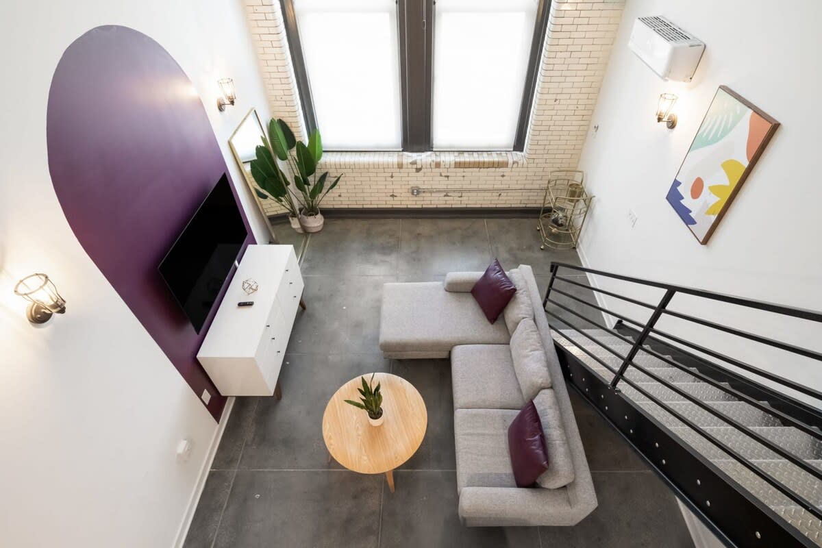 An aerial view of the living room showcasing a grey sectional sofa, a round wooden coffee table, and a white dresser with a TV on a purple accent wall. A black metal staircase leads to an upper level.