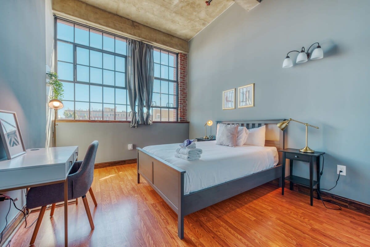 A bright bedroom with light blue walls, a dark grey bed frame, and a white desk with a grey chair. Large industrial-style windows with dark drapes overlook an urban landscape, and the room features warm-toned hardwood floors.