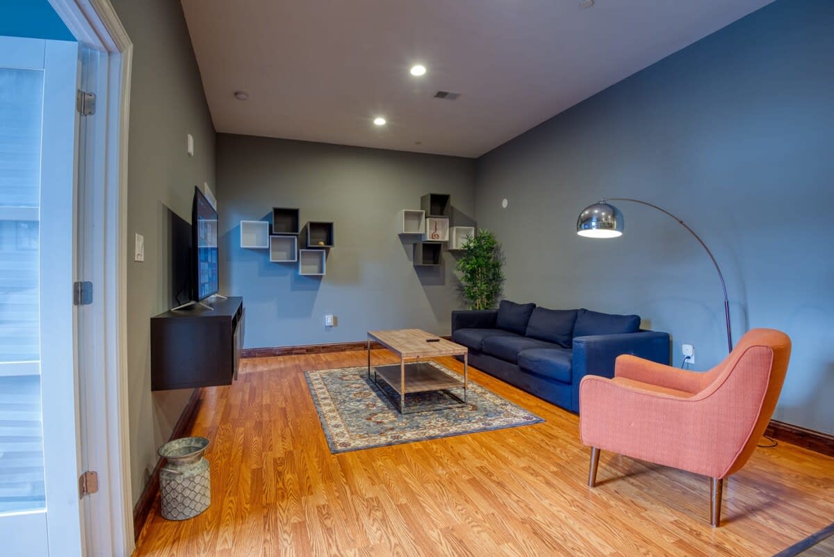A wide view of the living room showcasing light hardwood floors, dark gray walls, a dark blue sofa, and an orange armchair. A wooden coffee table is centered on a patterned rug, and a large arc floor lamp provides additional lighting, with decorative cube shelves on the wall.