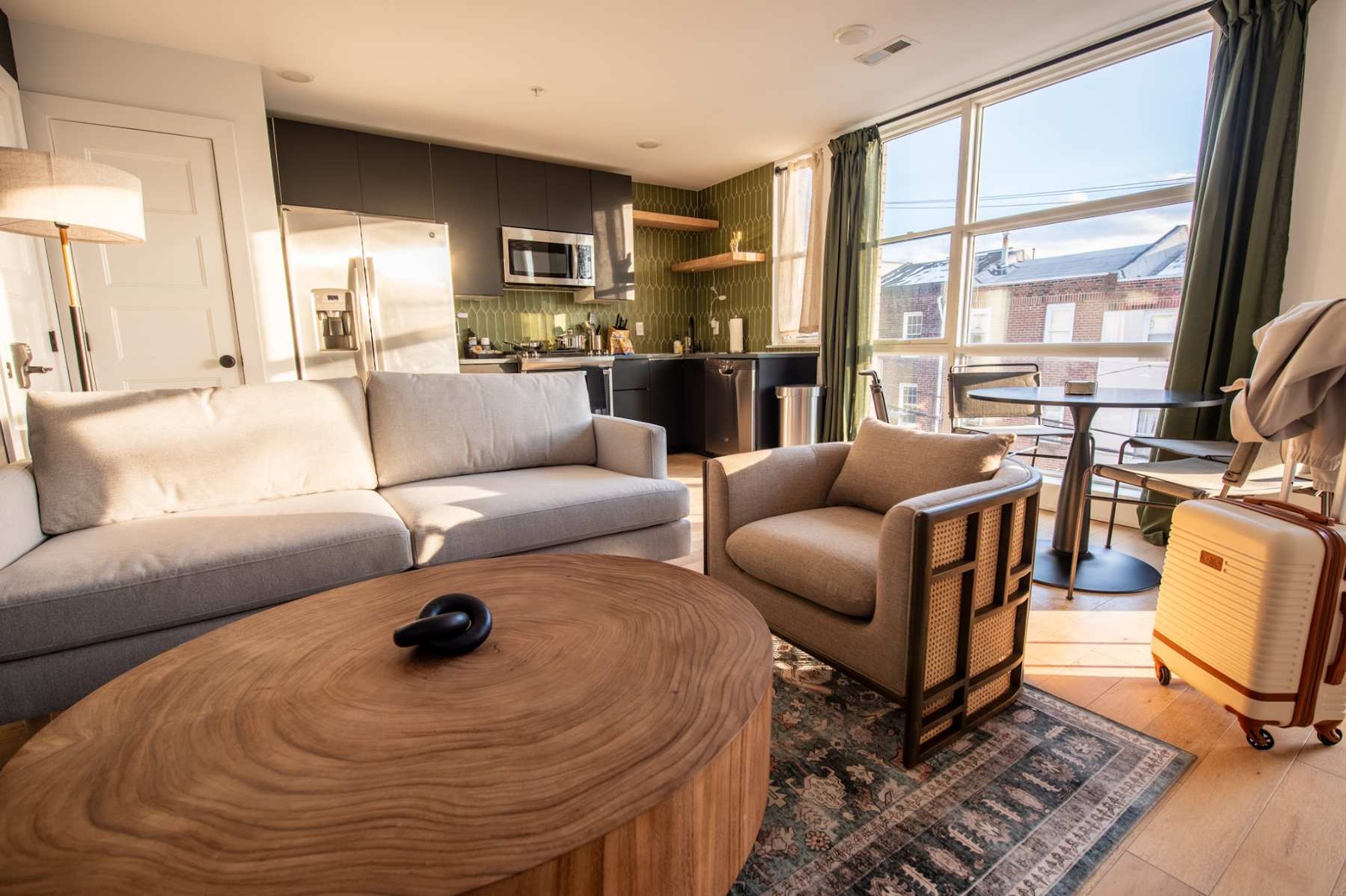 An inviting open-concept living room with a light grey sofa, a matching armchair with rattan detailing, and a large, organic-shaped wooden coffee table centered on an area rug. The space is brightly lit by a large window, with the kitchen and dining area visible in the background.