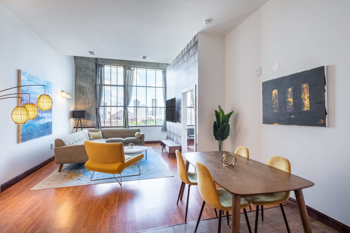 Open-concept living and dining area with a light-colored sectional sofa, a yellow accent chair, a wooden dining table with yellow chairs, and large windows overlooking a city skyline.