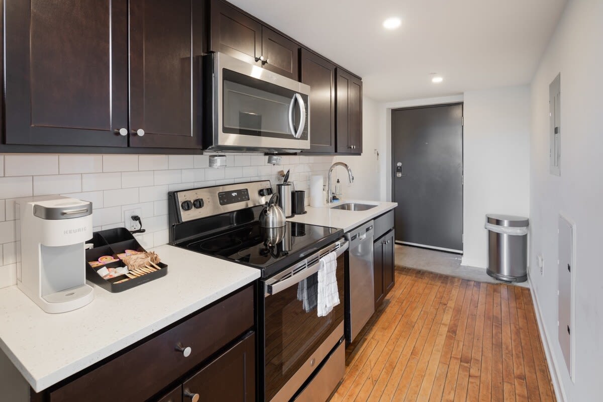 A modern kitchen with dark wood cabinetry, white subway tile backsplash, and stainless steel appliances including a microwave, stove, and dishwasher, alongside a white Keurig coffee maker on the counter.